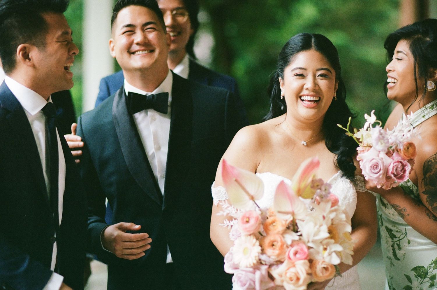 Radiant bride laughs with wedding party while holding pink bouquet at outdoor ceremony.