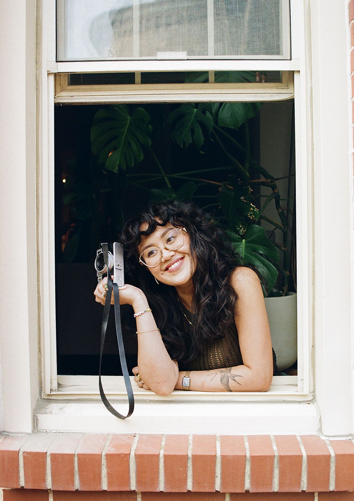 Smiling woman with curly hair and glasses holds a vintage camera while leaning out of an open window with plants behind her.