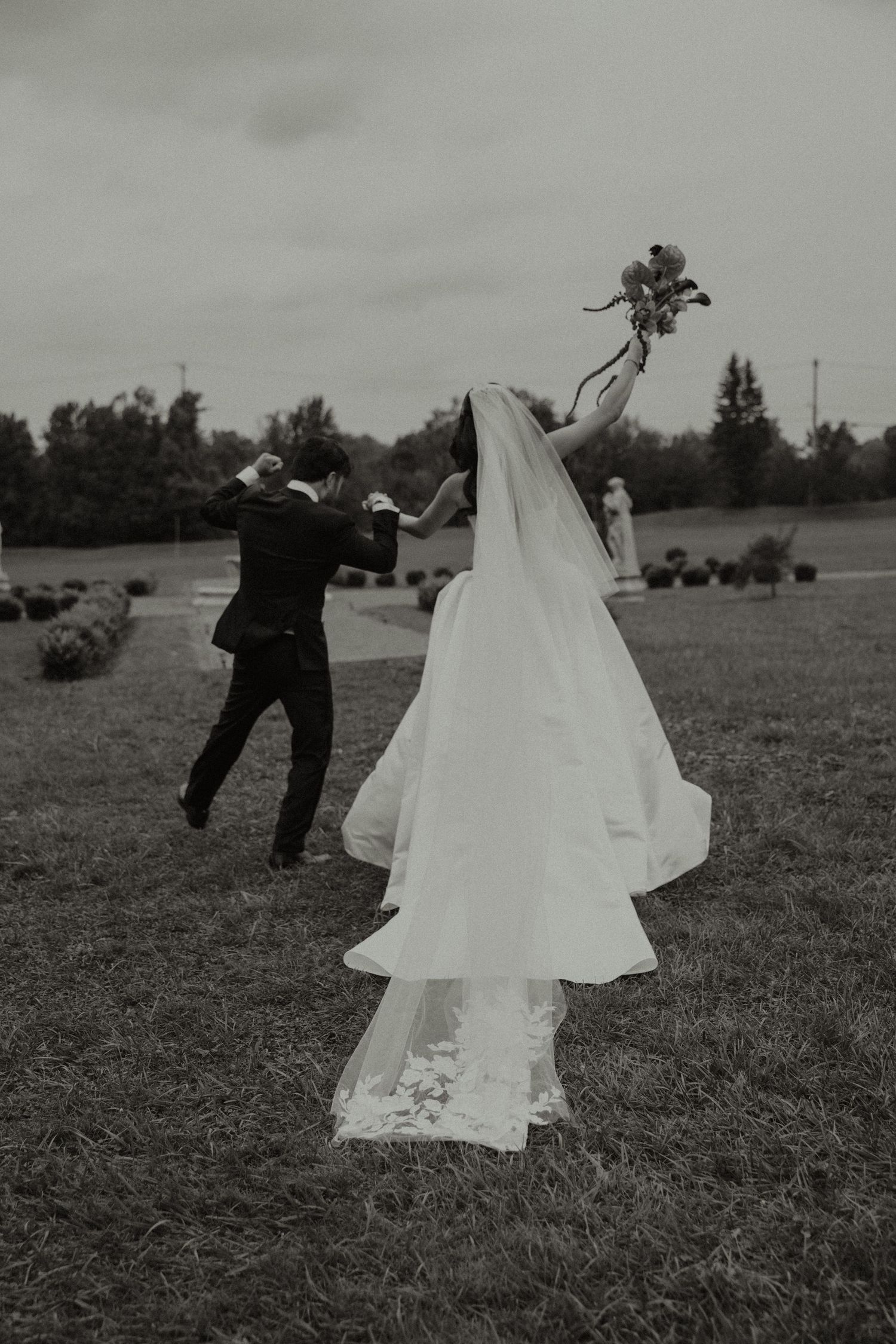 Black and white photo of bride and groom dancing joyfully outdoors, bride holding bouquet aloft, veil flowing in the wind.