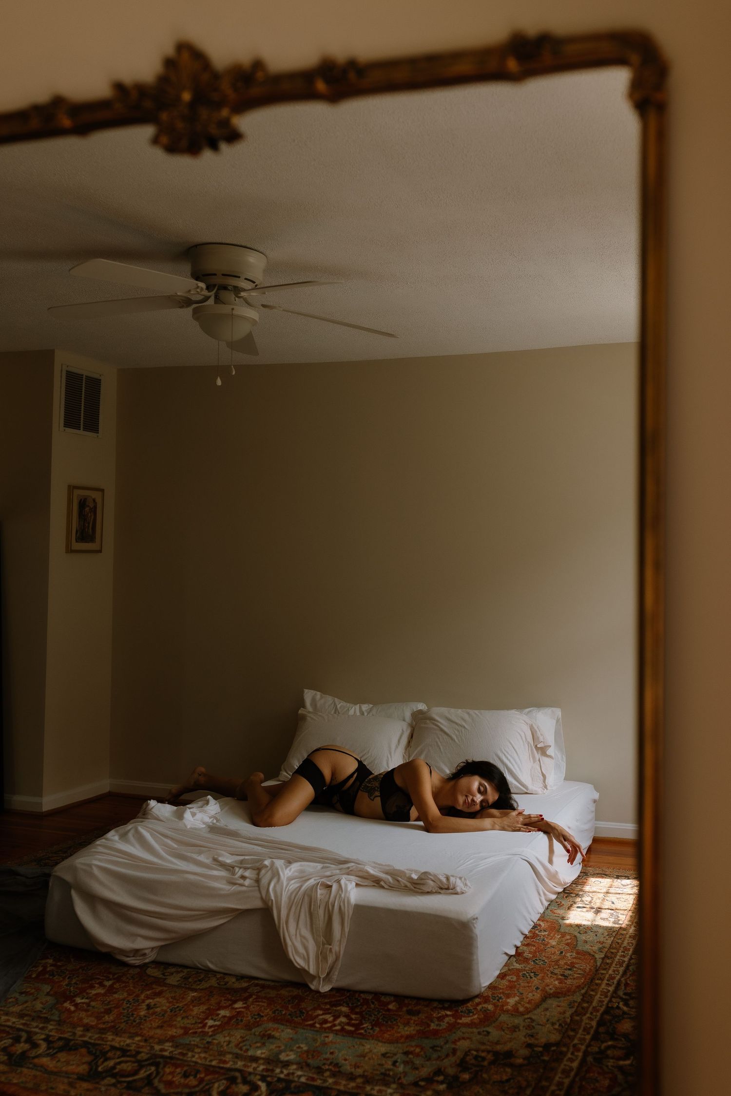 Woman in black lingerie reclining on white bed, reflected in ornate gold mirror in cozy bedroom.