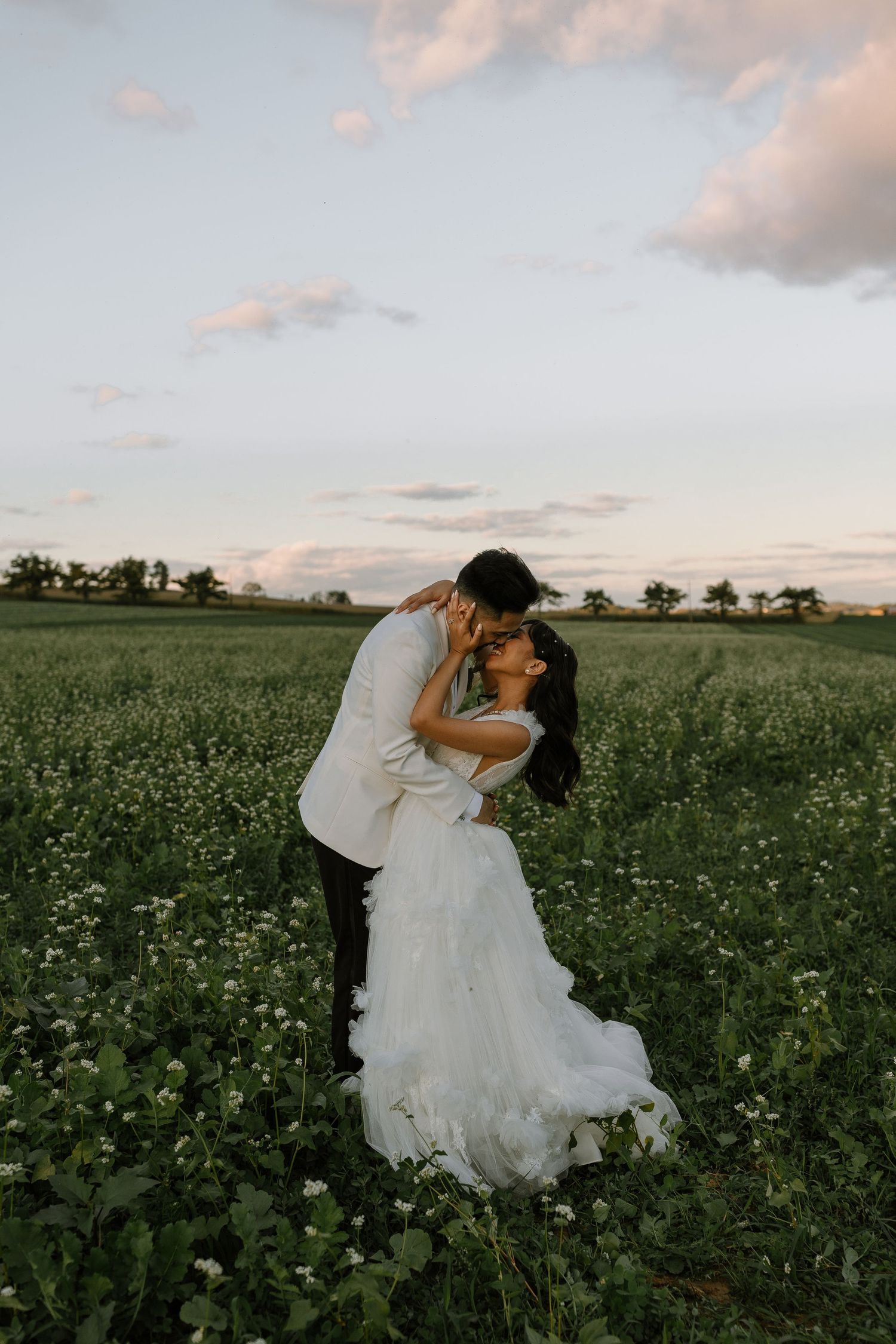 A bride and groom share a romantic kiss in a lush green flower field at sunset during their wedding day.