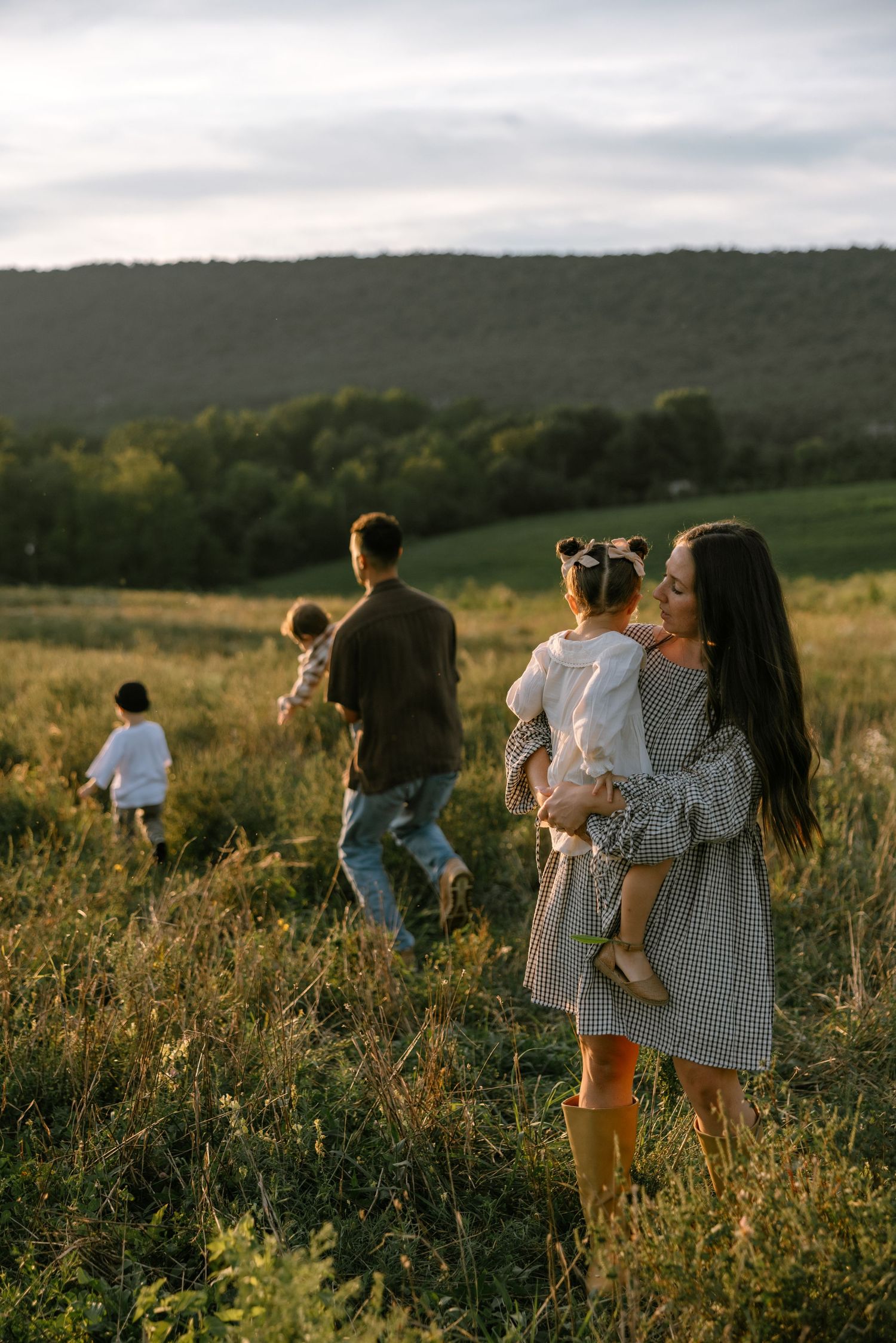 Family walking in a sunlit meadow with green hills, mother holding daughter, father carrying toddler at golden hour.