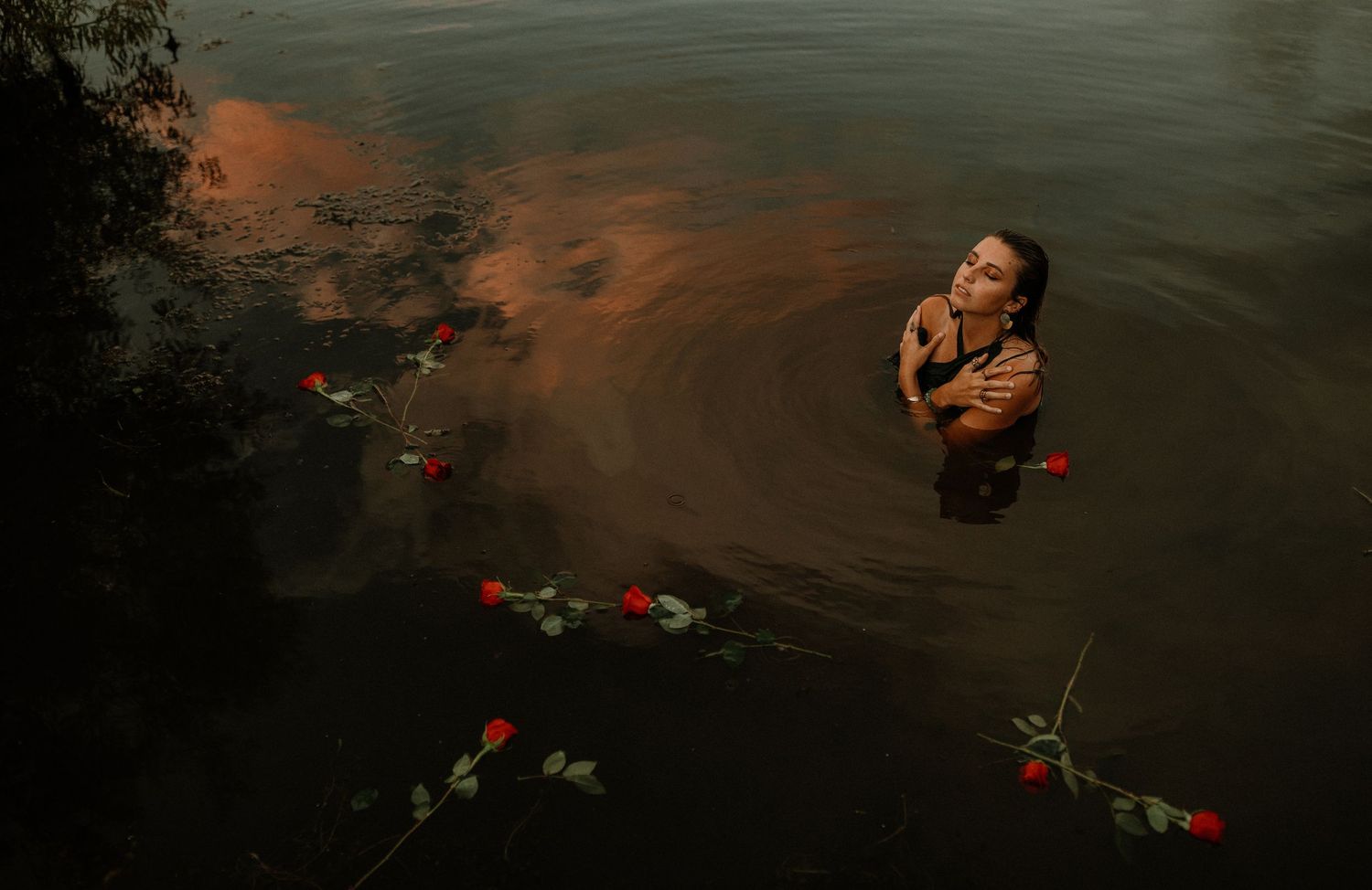A woman stands waist-deep in dark water surrounded by floating red roses at dusk.