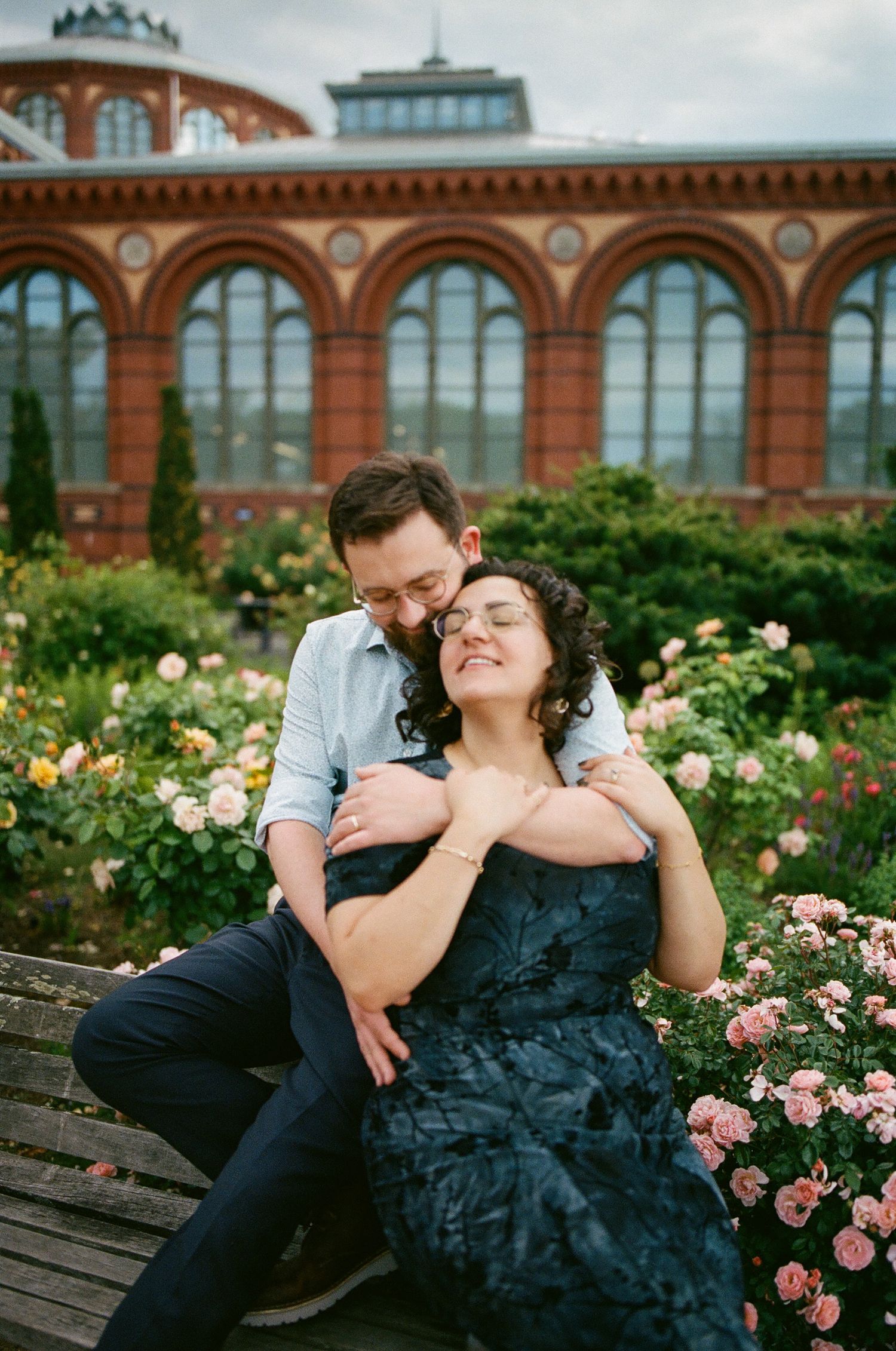 Couple embracing on a bench in a rose garden, with a grand brick building with arched windows behind them.