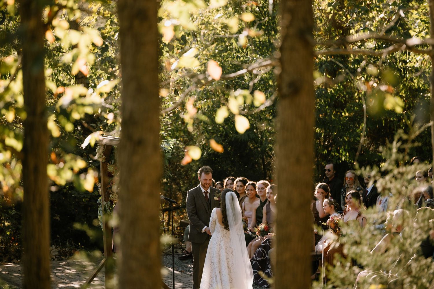 Outdoor wedding ceremony in a lush forest setting with bride and groom at the altar surrounded by tall trees.