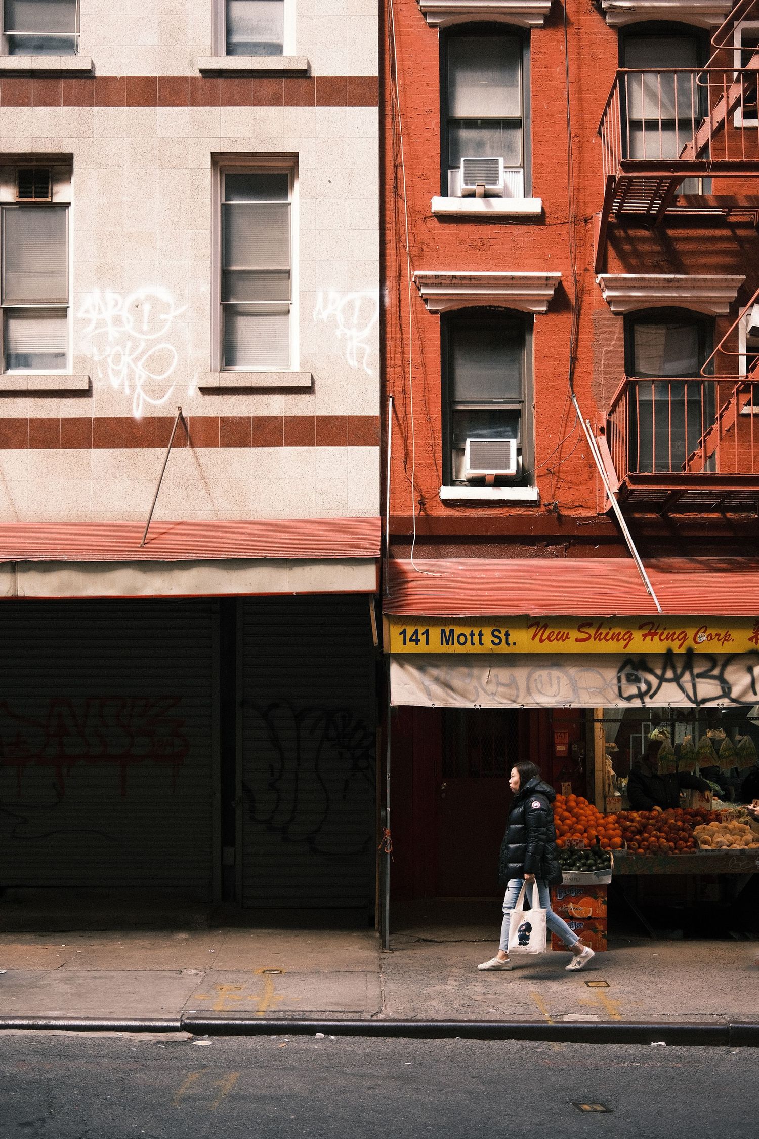 Person walking past 141 Mott St in NYC, with red brick buildings, fire escapes, graffiti, and a fruit stand.