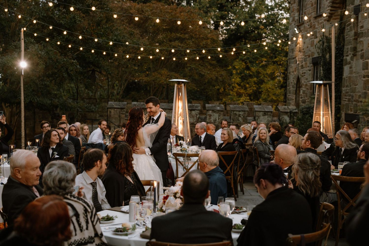 Couple shares first dance at elegant outdoor wedding reception with string lights and guests seated at decorated tables.