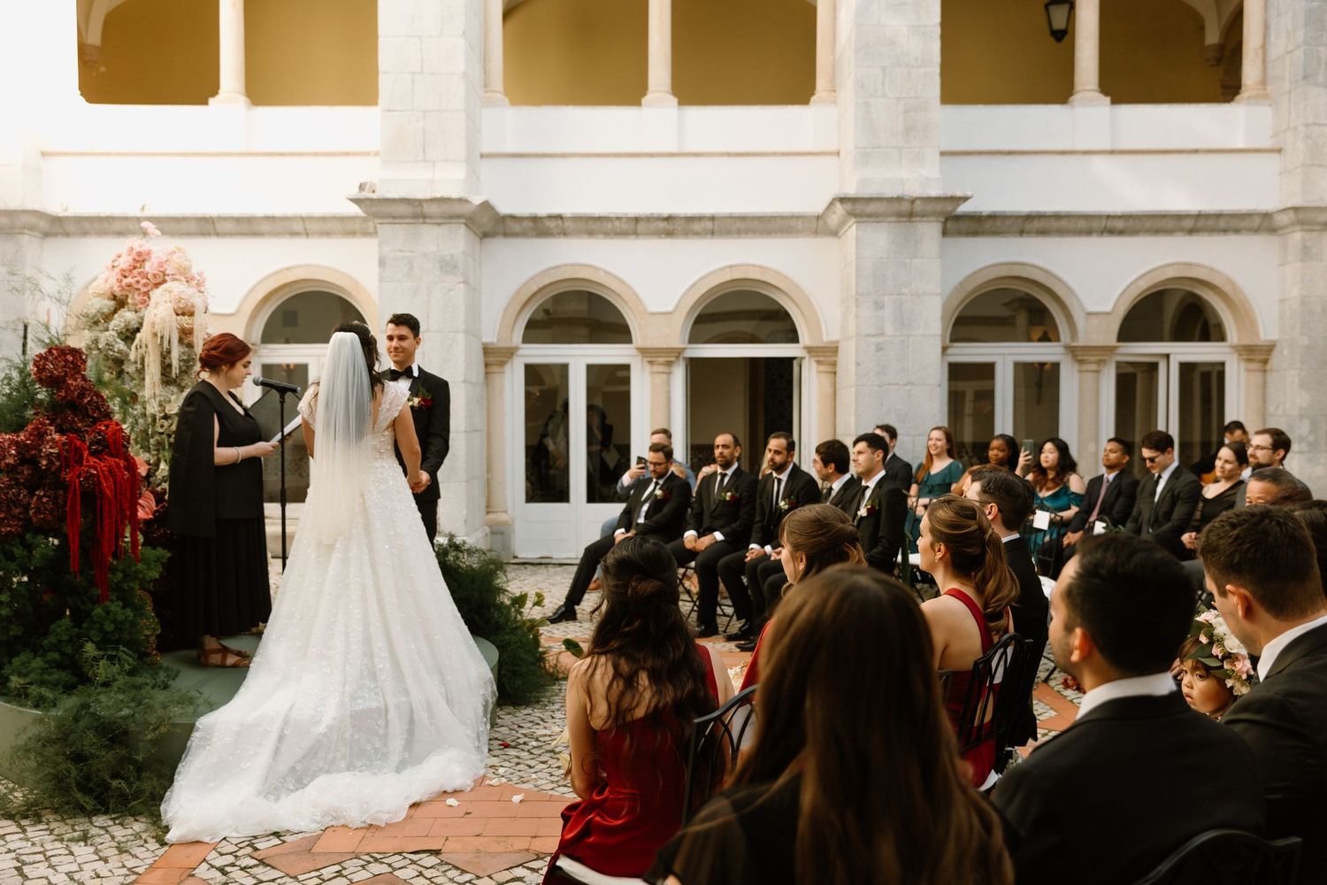 Elegant outdoor wedding ceremony in a courtyard with bride and groom at altar, guests seated, floral arrangements, and arched architecture.