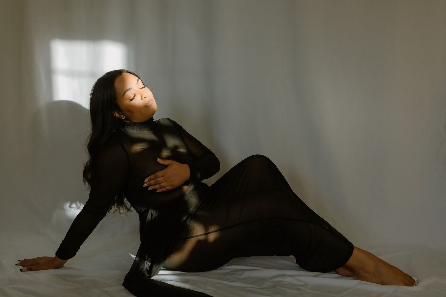 Pregnant woman in black sheer dress poses gracefully on floor in soft natural light for maternity photography session.