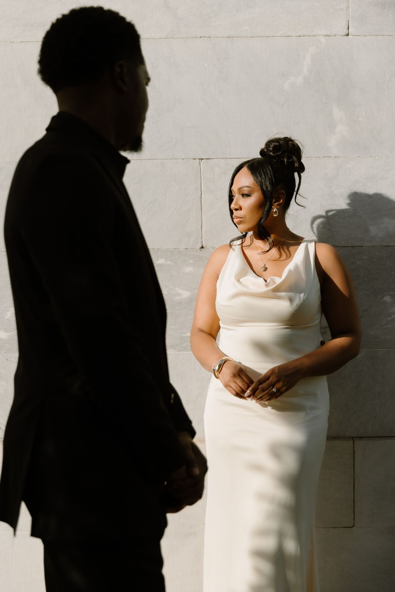 A bride in a white satin gown stands before a stone wall, facing a groom in a black suit in soft sunlight.