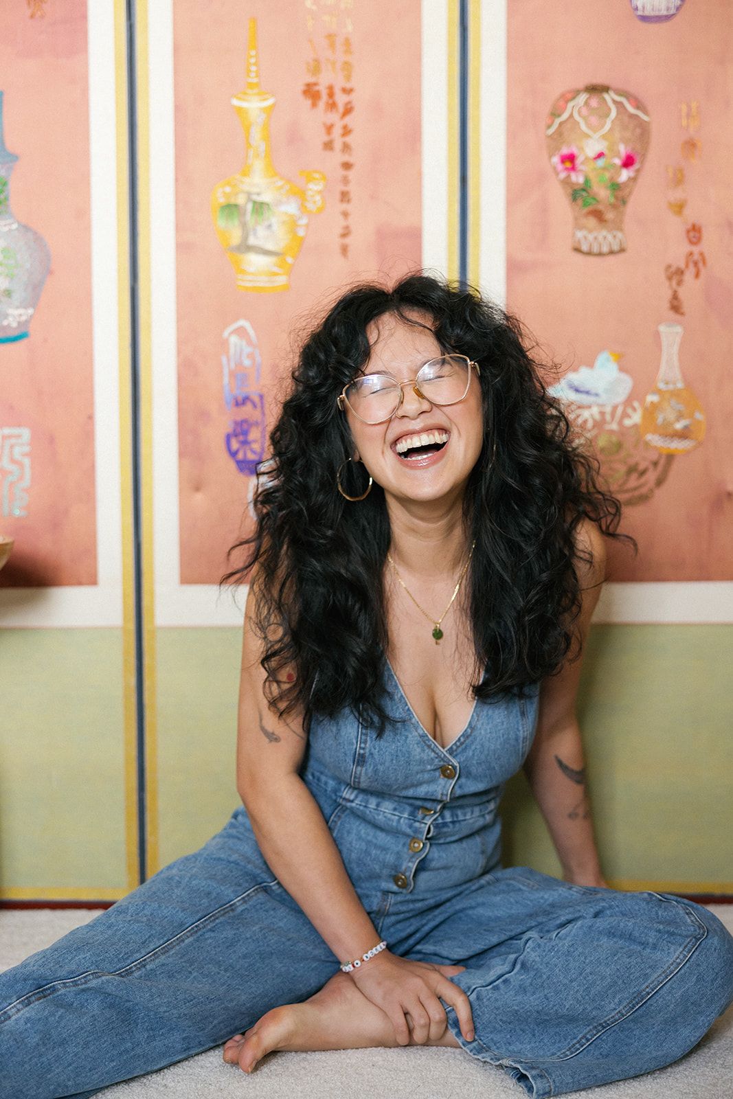 Smiling woman with long curly black hair and glasses sits laughing in a denim jumpsuit before a folding screen painted with vases.