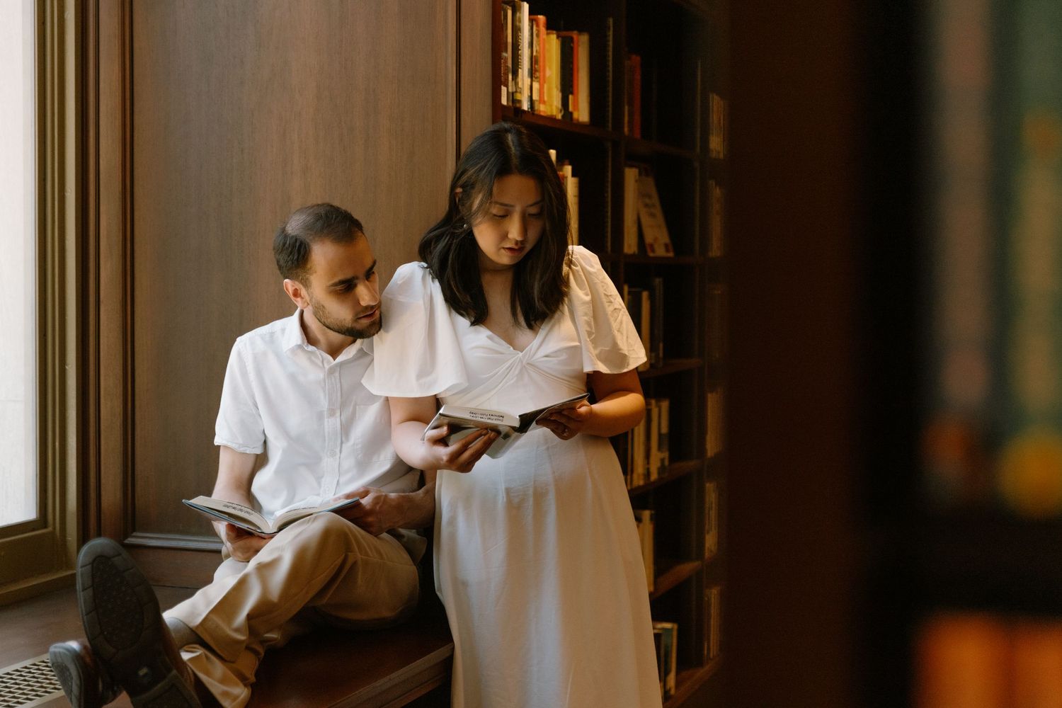 A man and woman in a cozy library reading a book together, she wears a white dress near wooden bookshelves.