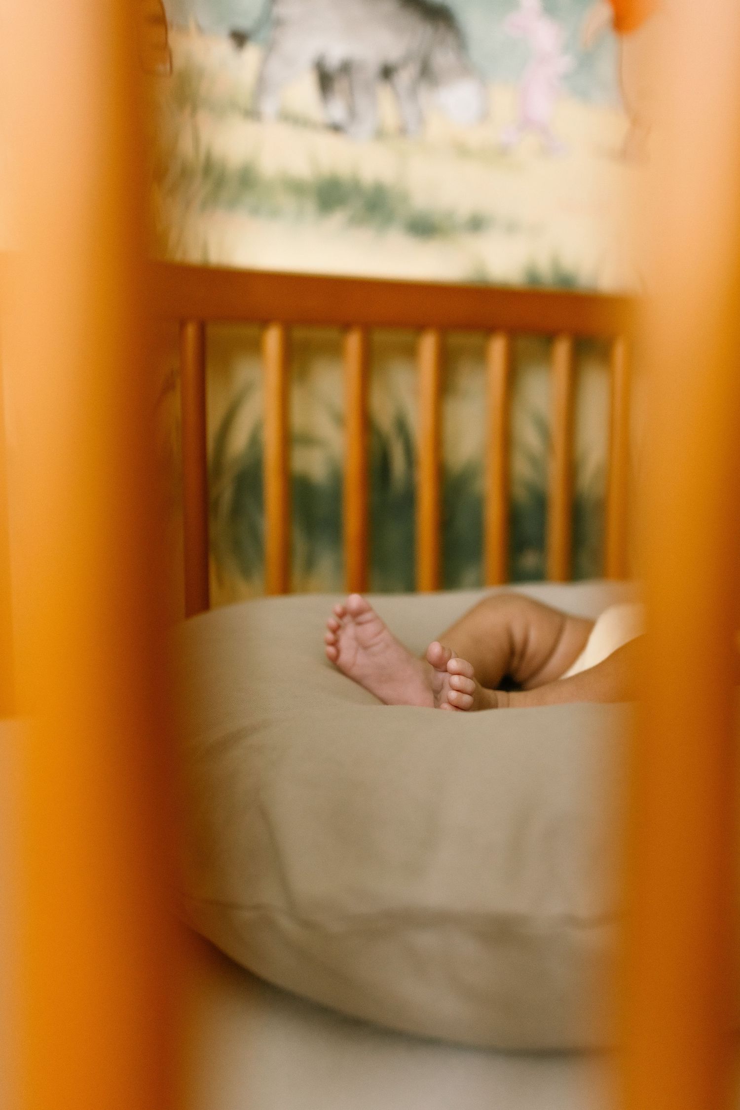 Tiny baby feet peeking out from under a blanket in a wooden crib with animal-themed nursery decor.