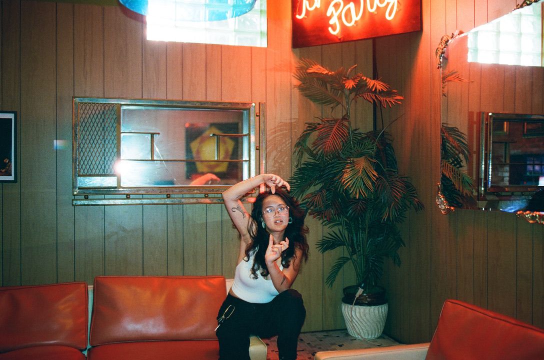 Person posing in retro diner with wood paneling, red leather couch, neon sign, and tropical plant.