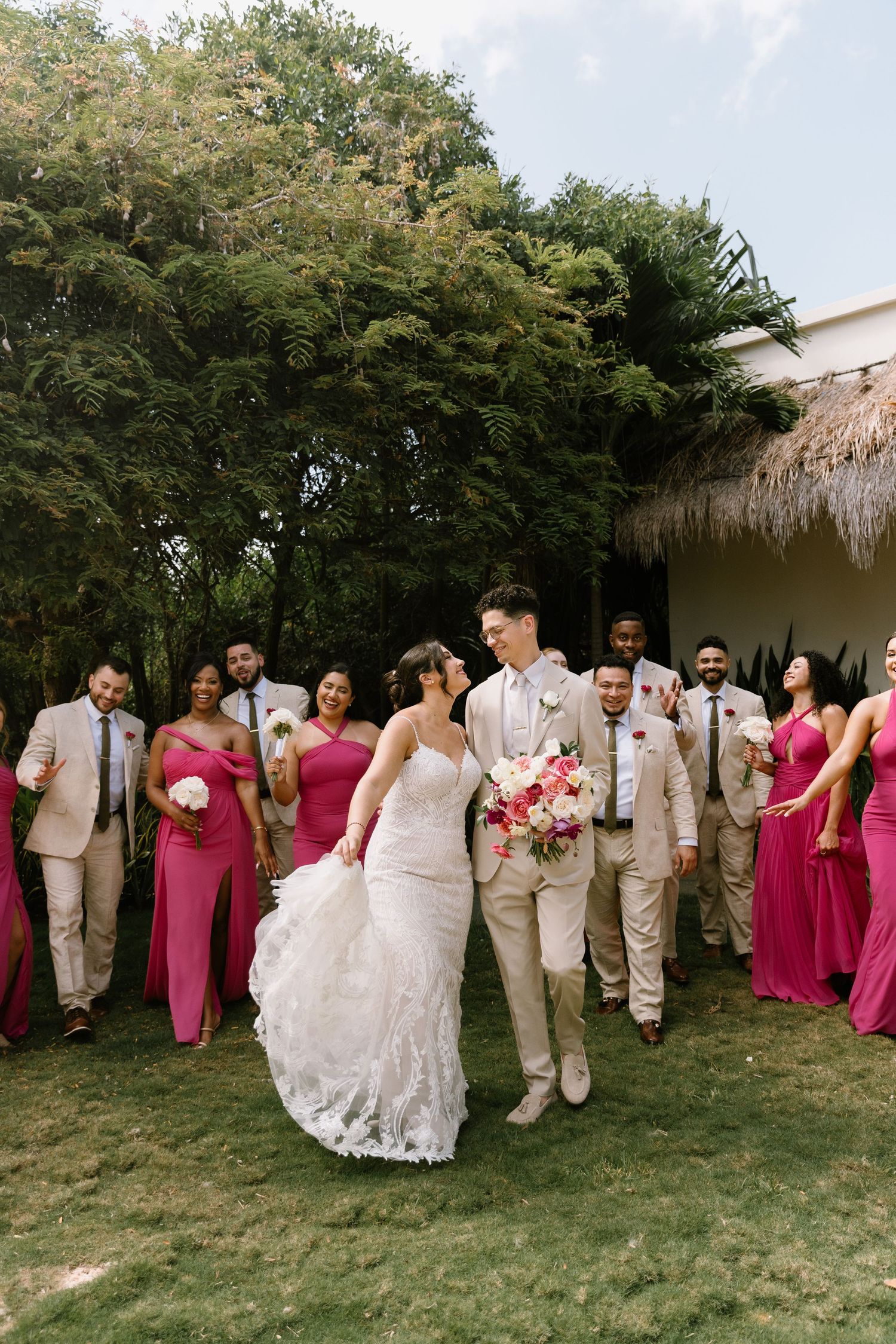 Bride and groom walk together as wedding party in pink dresses and tan suits celebrate outdoors amid lush tropical greenery.