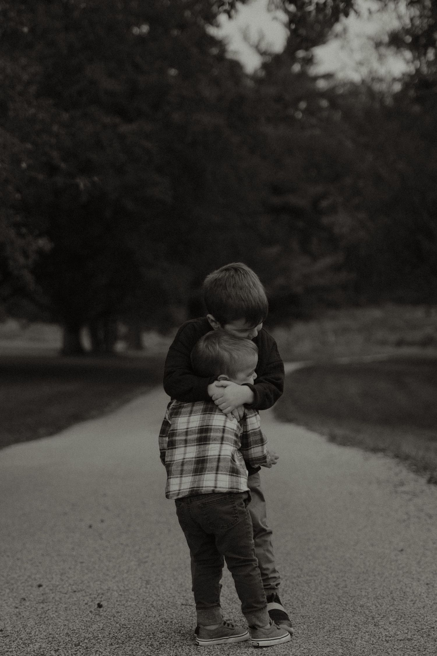 Two young boys share a tender hug on a tree-lined path in a black and white outdoor portrait.