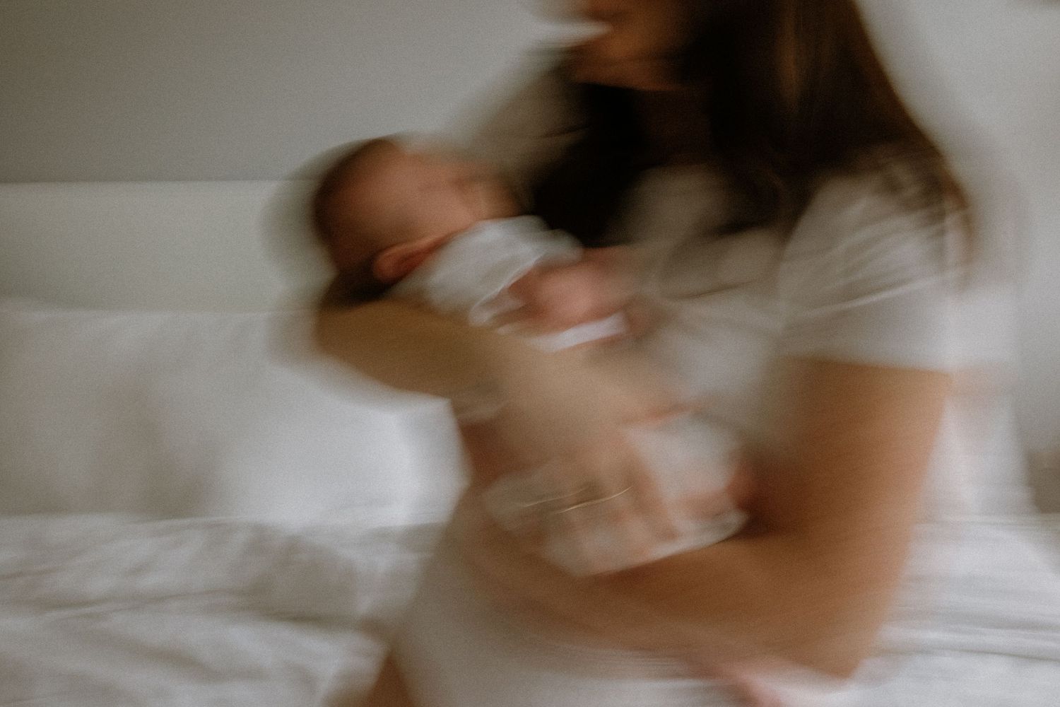 Blurry motion photo of a mother in white dress cradling her newborn baby while sitting on a white bed.