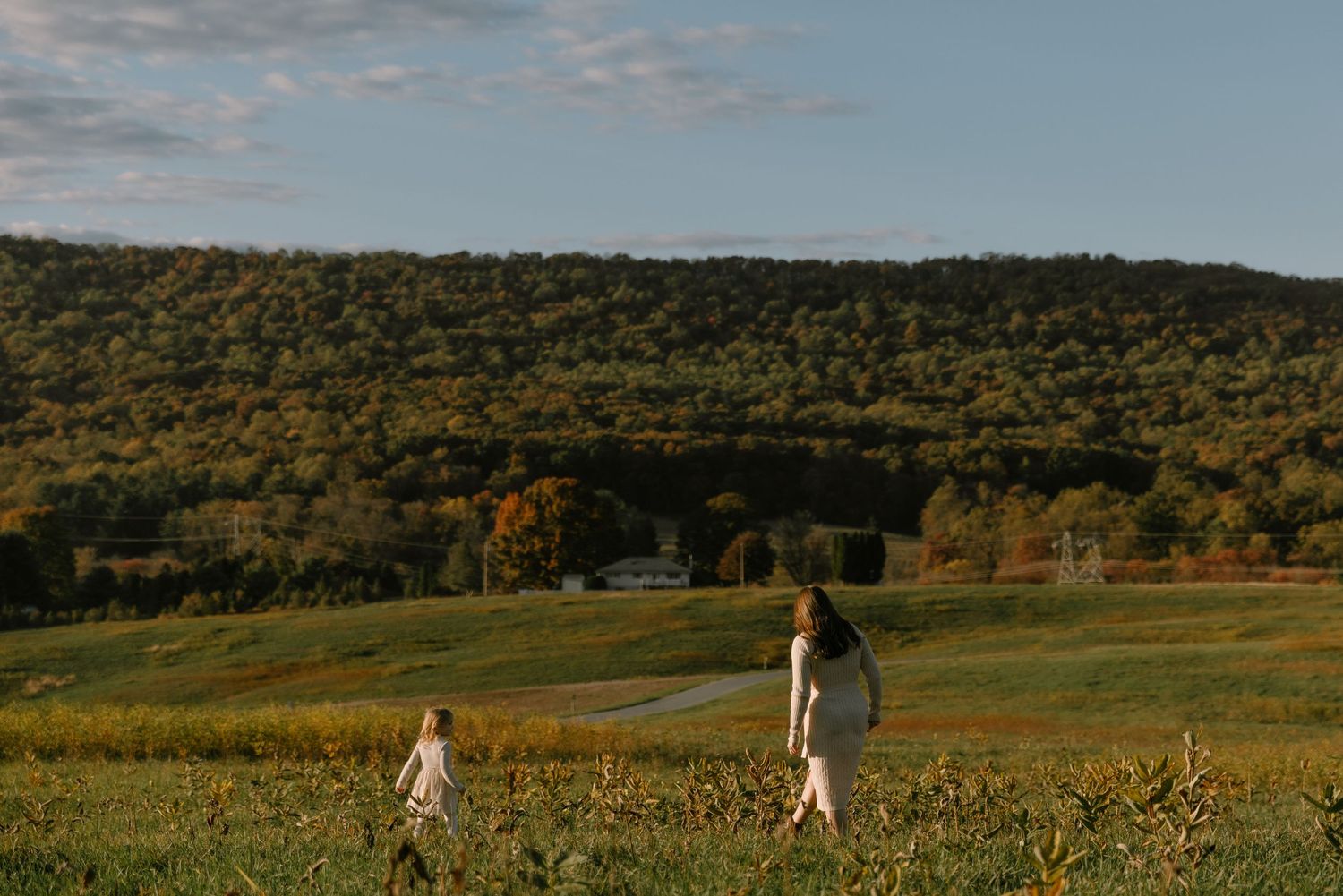 A mother and child walk through a lush green field with autumn hills and a farmhouse in the background.