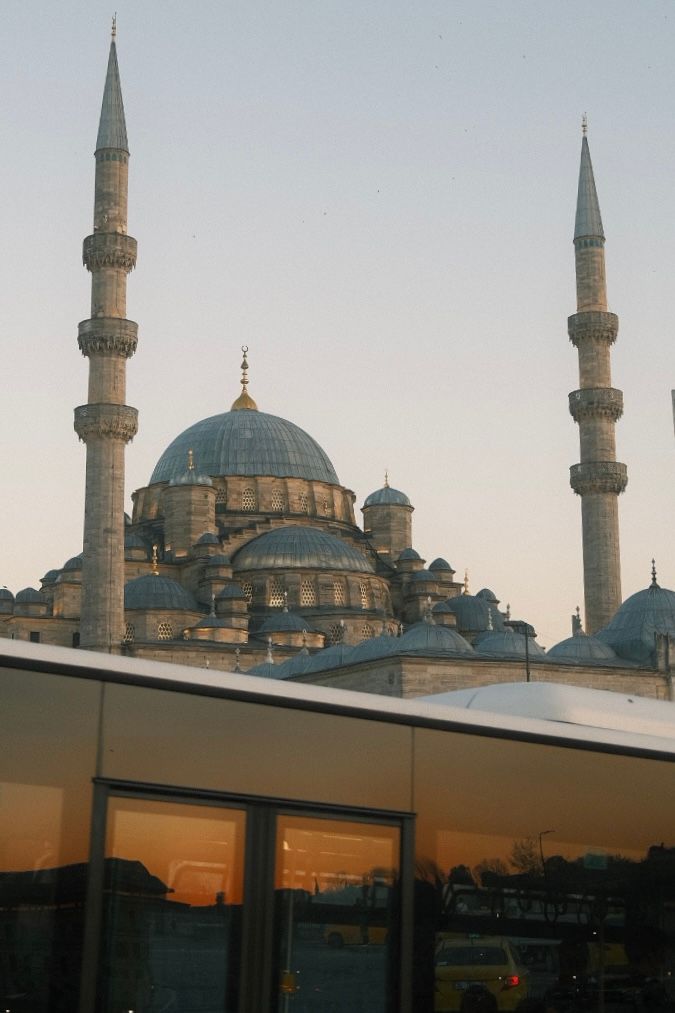 The New Mosque in Istanbul, Turkey, with its iconic domes and minarets reflected in glass at sunset.