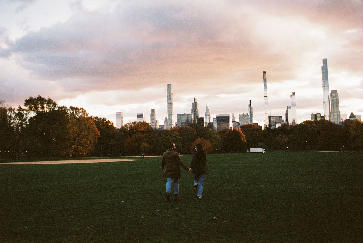 Two people walk hand-in-hand across Central Park's Great Lawn at sunset with NYC skyline in the background.