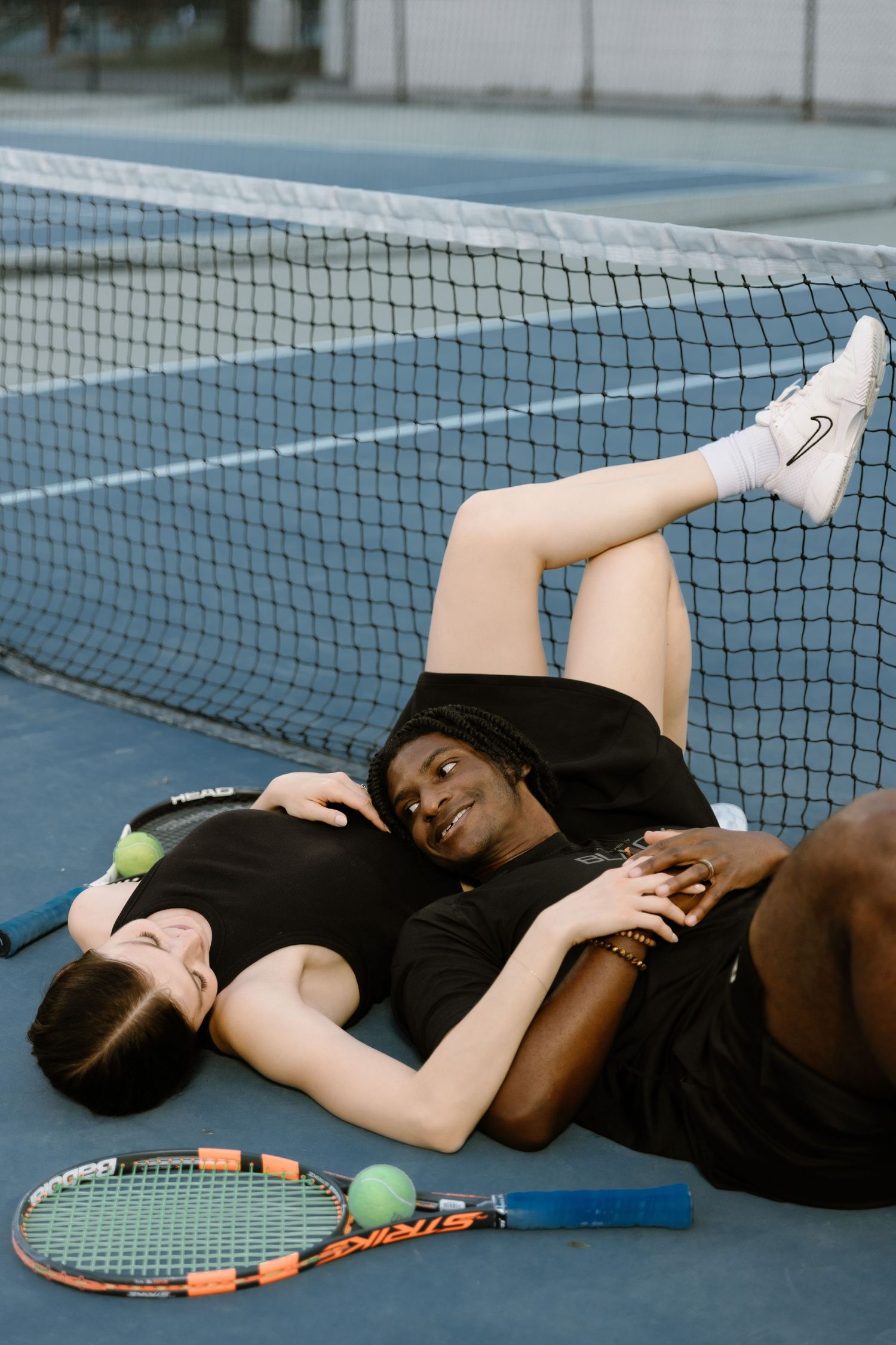 Two tennis players resting together on a blue court beside the net with rackets and balls nearby.