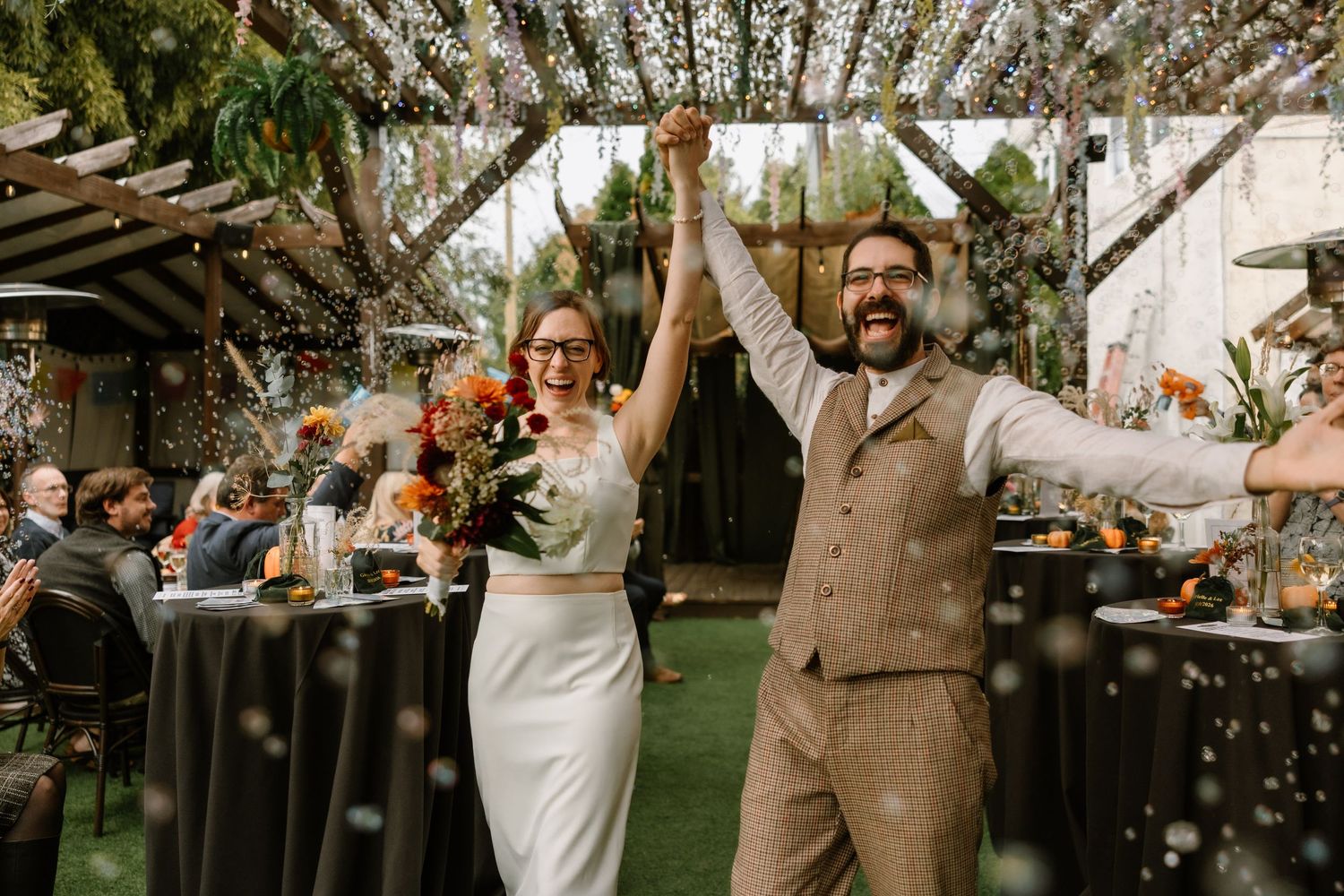 Joyful newlyweds celebrate their wedding reception outdoors, raising hands under a floral pergola with colorful bouquet.