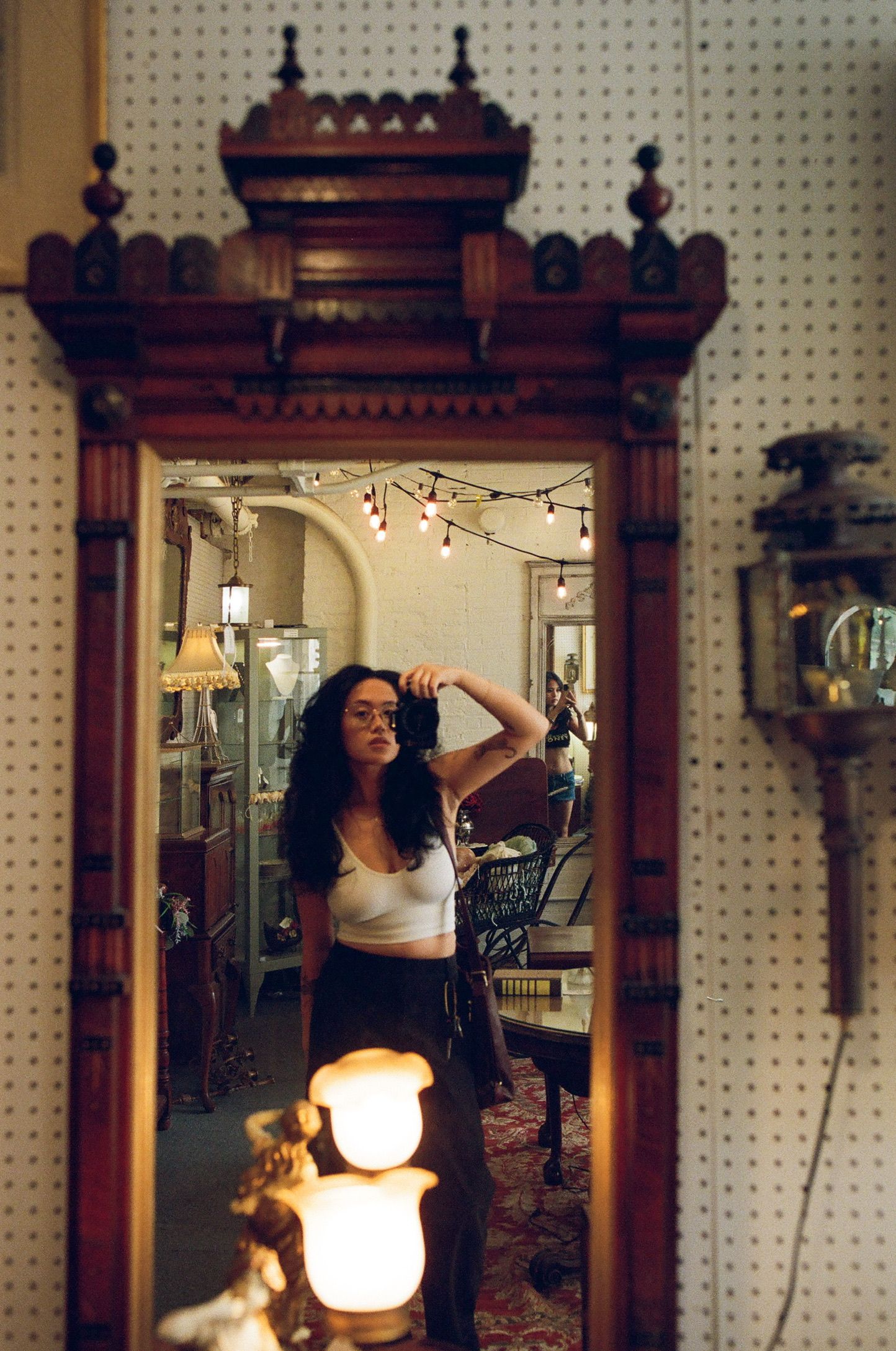 Woman taking mirror selfie in ornate antique wooden frame mirror surrounded by vintage lamps and string lights.