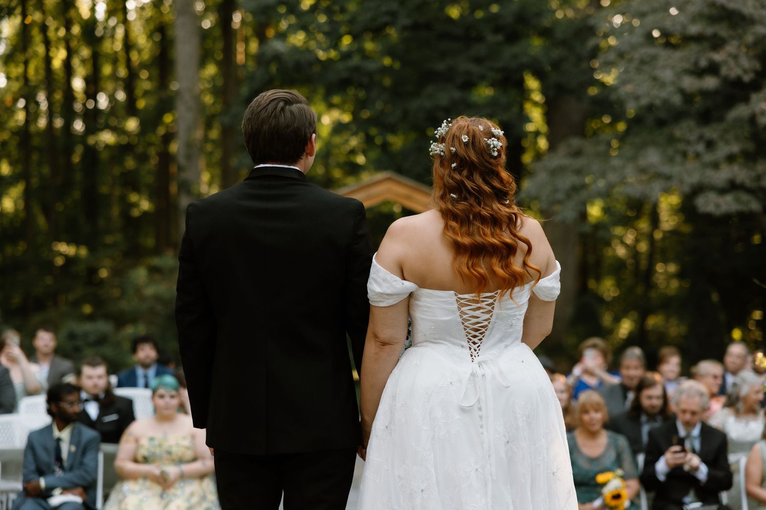 Bride and groom stand before outdoor wedding guests surrounded by lush green trees at a forest ceremony.