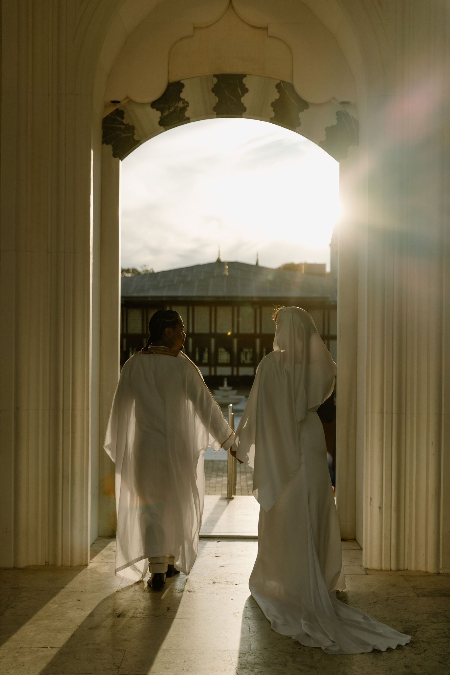 Two figures in white robes walk hand in hand through a sunlit archway at a mosque during golden hour.