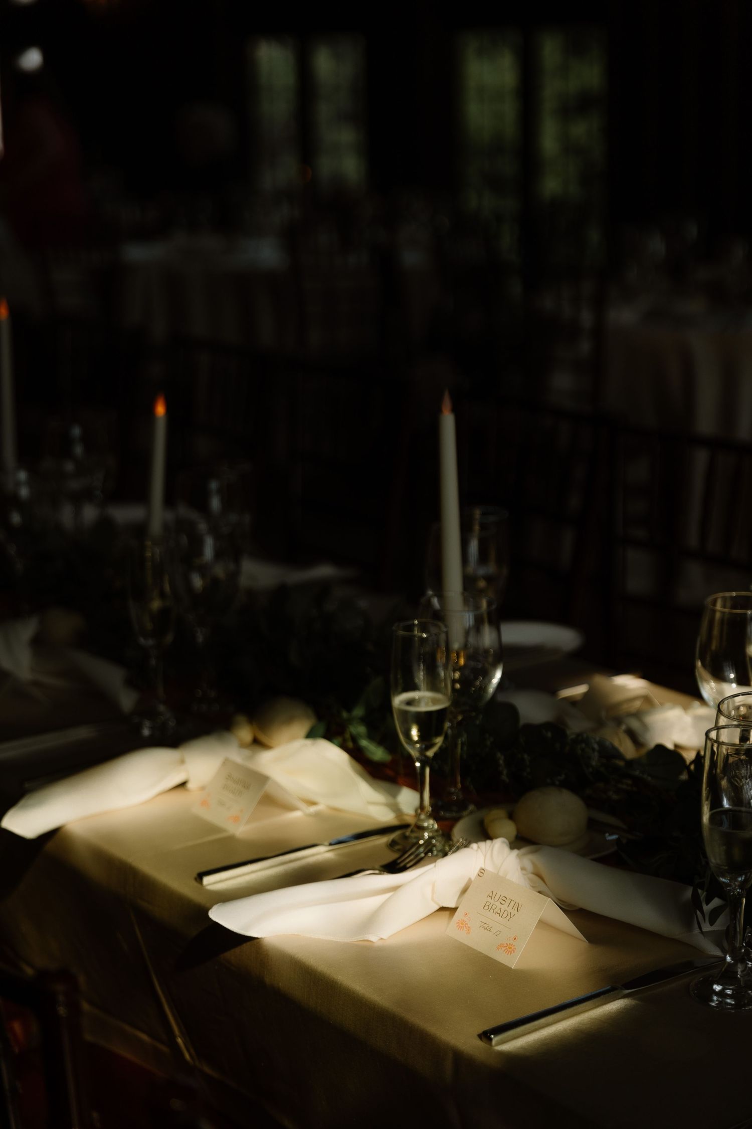 Elegant candlelit wedding reception table setting with white napkins, place cards, and greenery centerpiece.