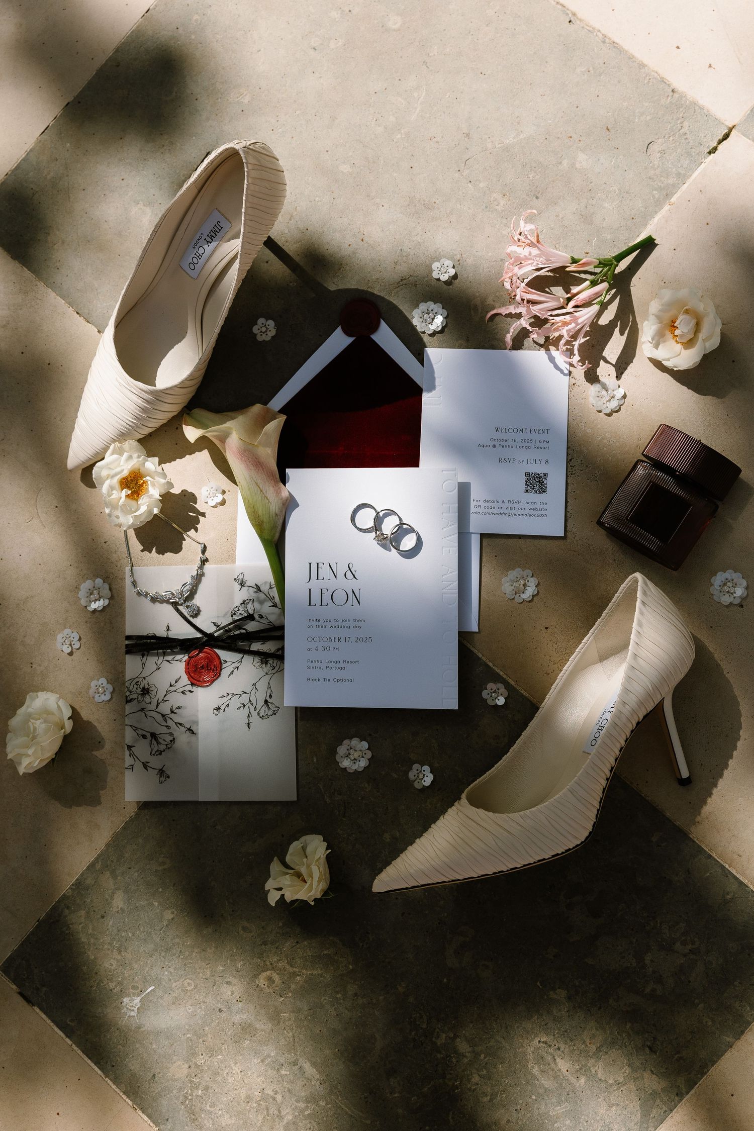 Elegant wedding flat lay with ivory heels, floral invitations, rings, and pink flowers on a stone surface.