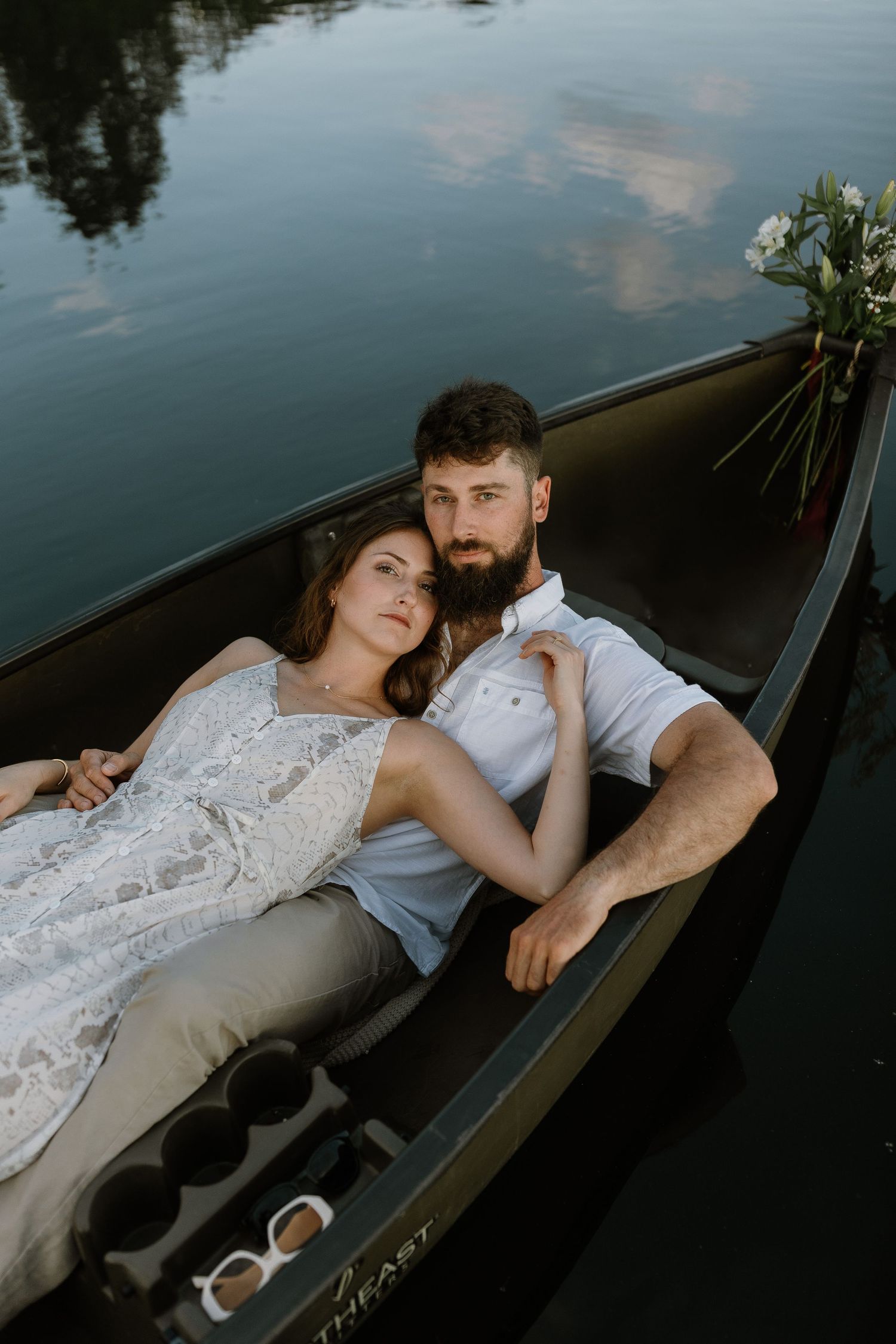 Romantic couple lying together in a gondola on calm water, woman in lace dress resting on bearded man's chest.