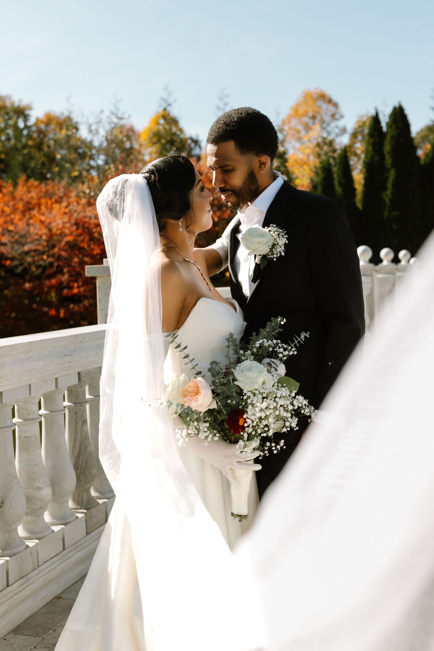 Bride and groom share a romantic moment on white marble stairs with autumn foliage backdrop, bride holding a bouquet.