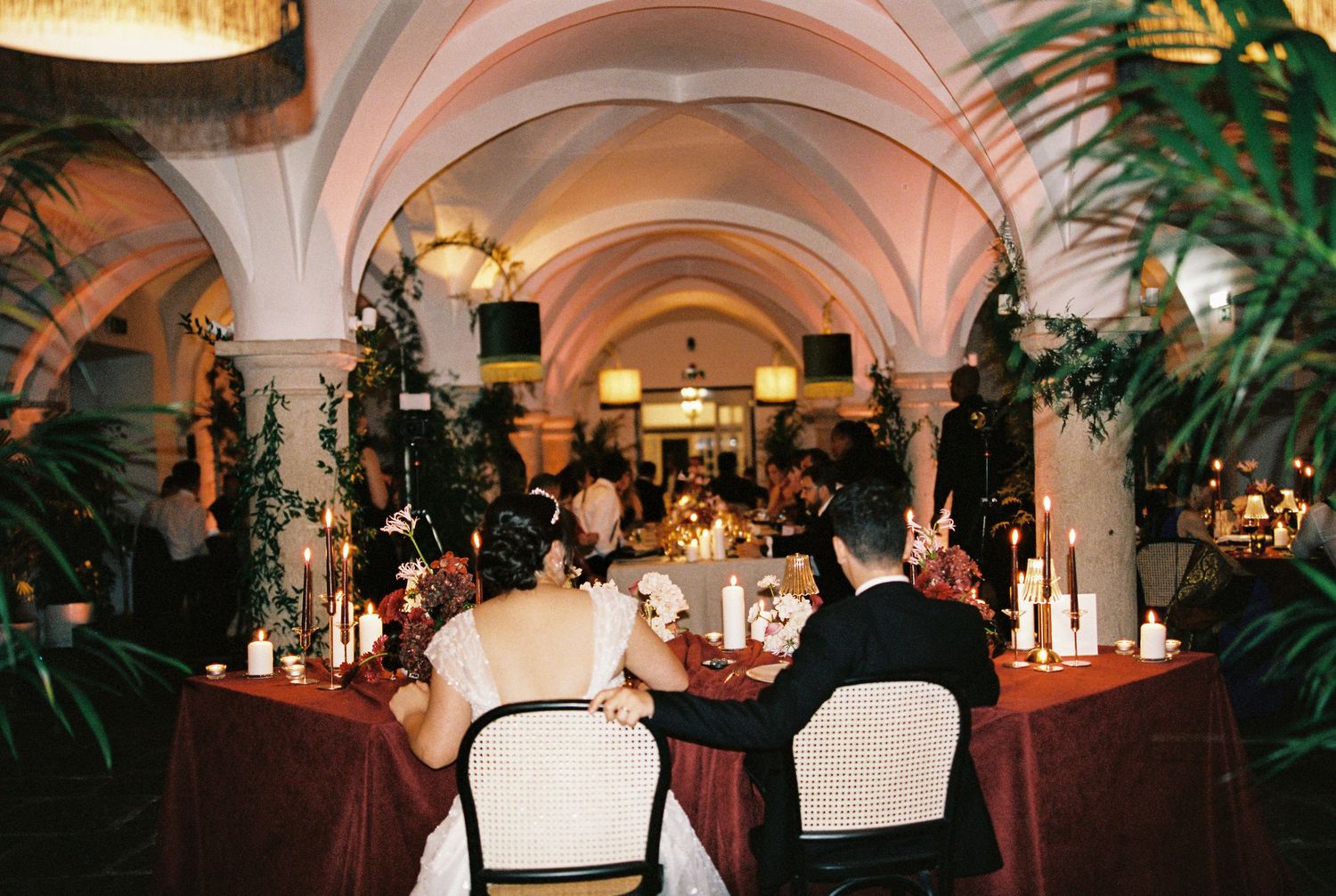 Elegant wedding reception in a vaulted venue with candlelit tables, lush greenery, and a couple seated at the sweetheart table.