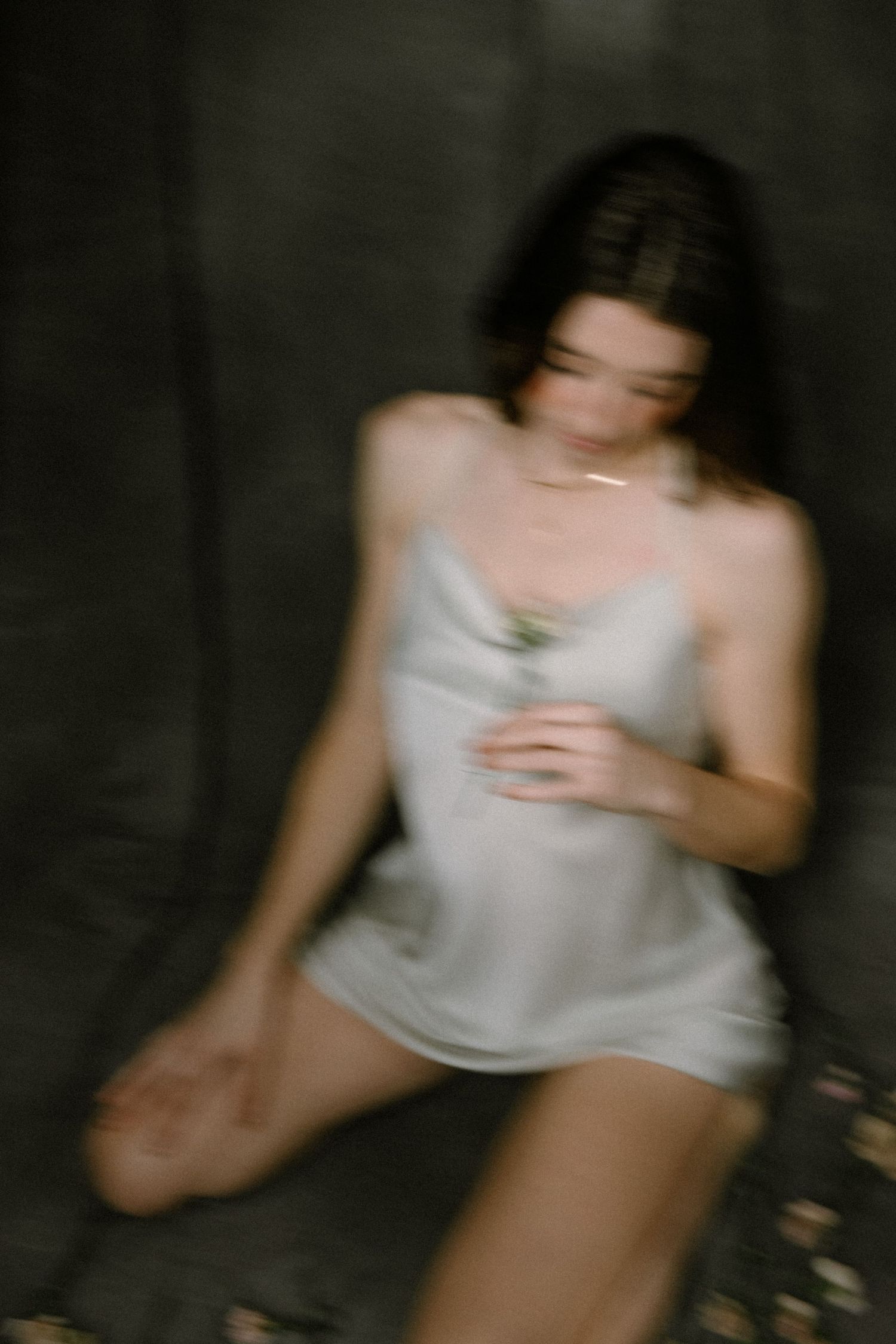 A blurred artistic photo of a young woman in a silver slip dress holding a flower against a dark background.