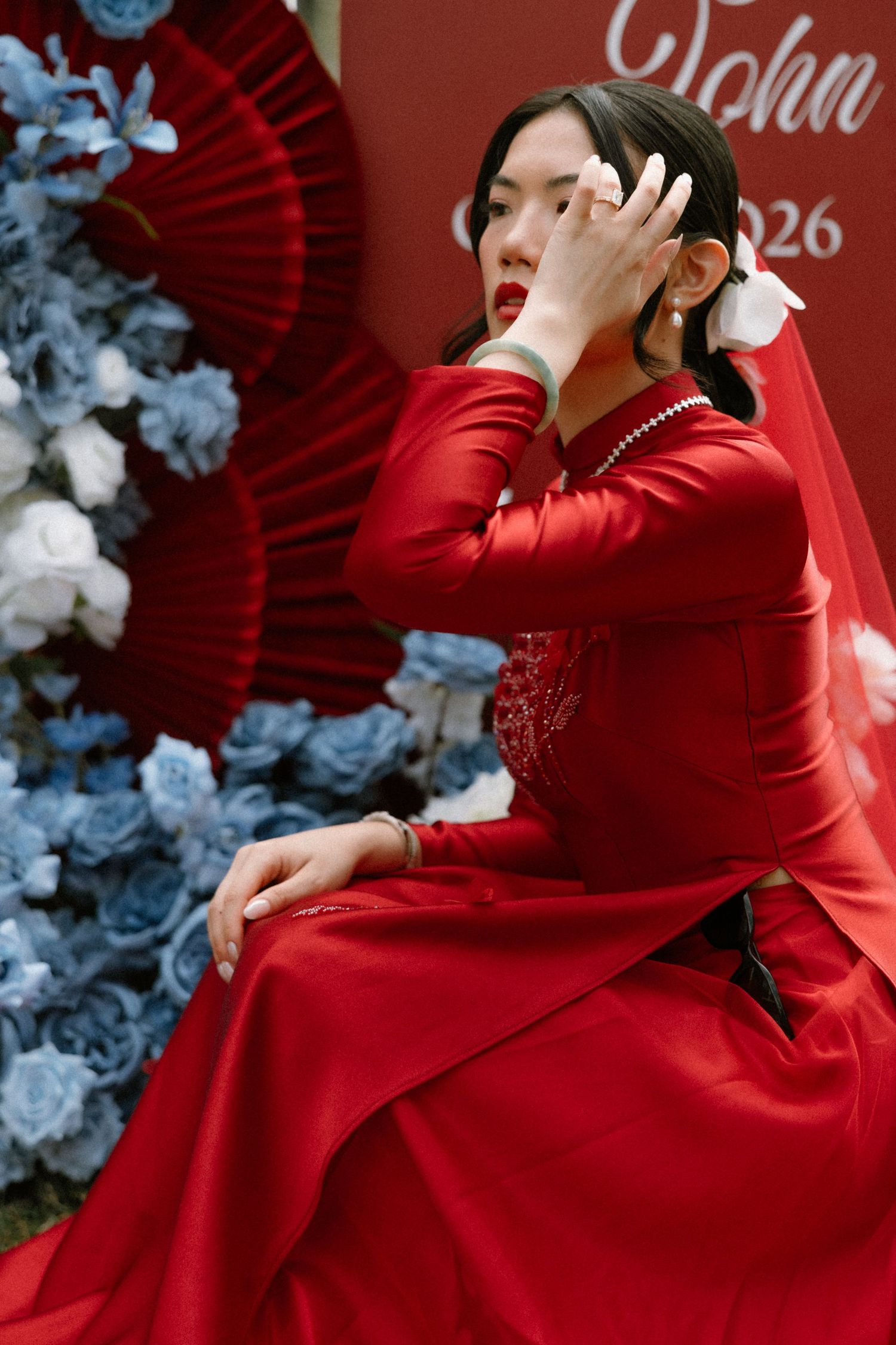 A woman in a red ao dai dress and veil sits gracefully among blue flowers and red fans at a wedding.