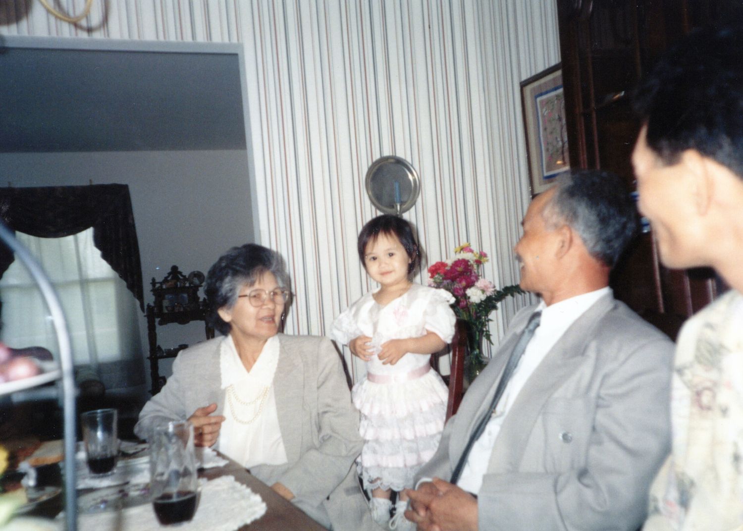 Elderly couple sitting with a young child in a white dress at an indoor family gathering.
