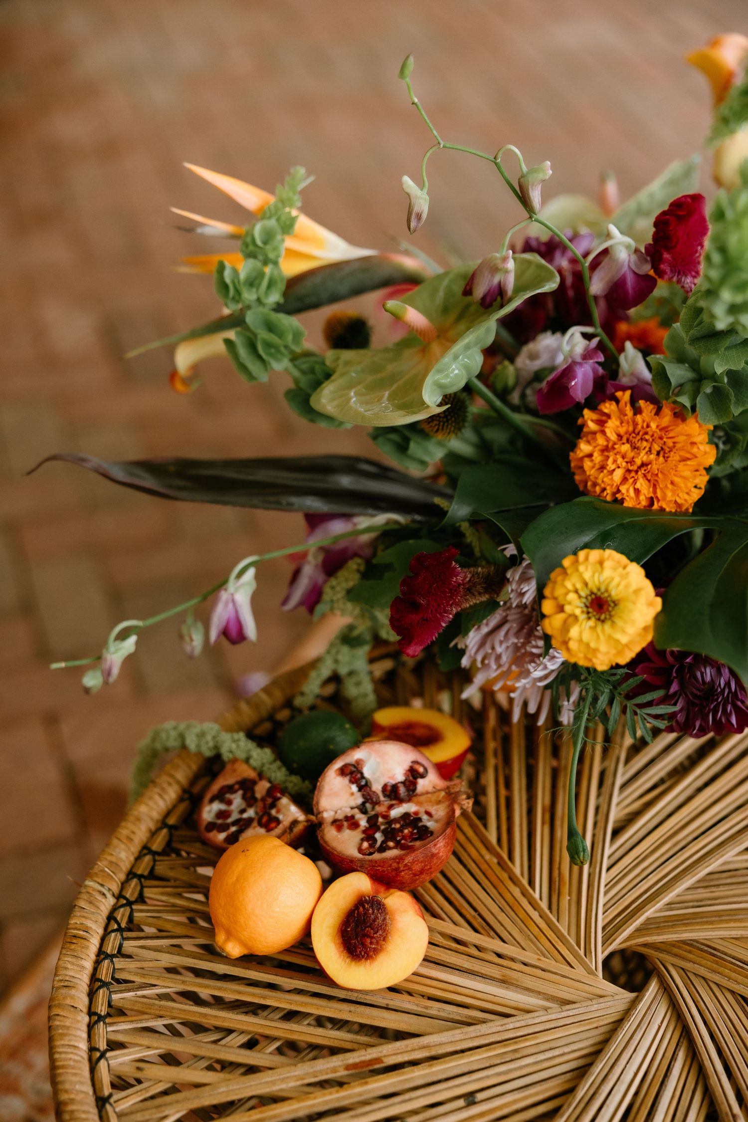 Vibrant floral arrangement with tropical flowers beside fresh peaches and pomegranate on a woven rattan table.