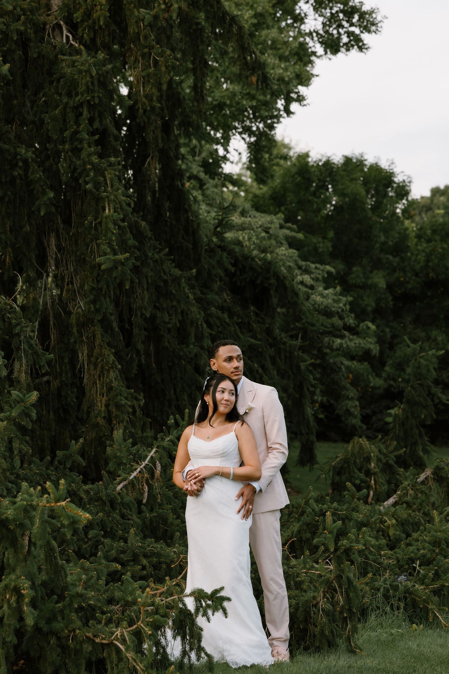 A bride and groom pose elegantly in front of lush green trees, the groom in a tan suit embracing the bride in a white gown.