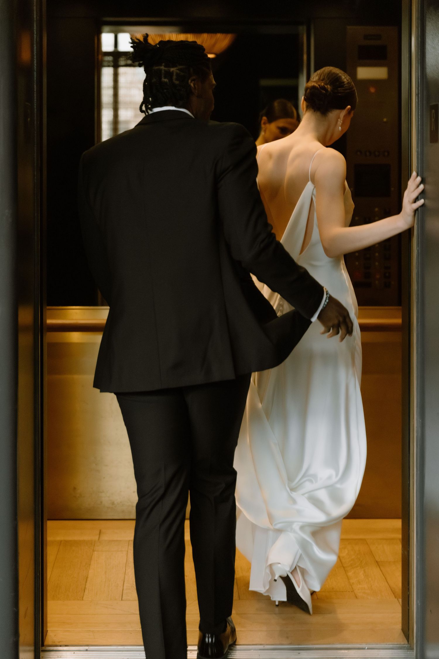 Bride in backless white gown and groom in black tuxedo entering an elevator together.