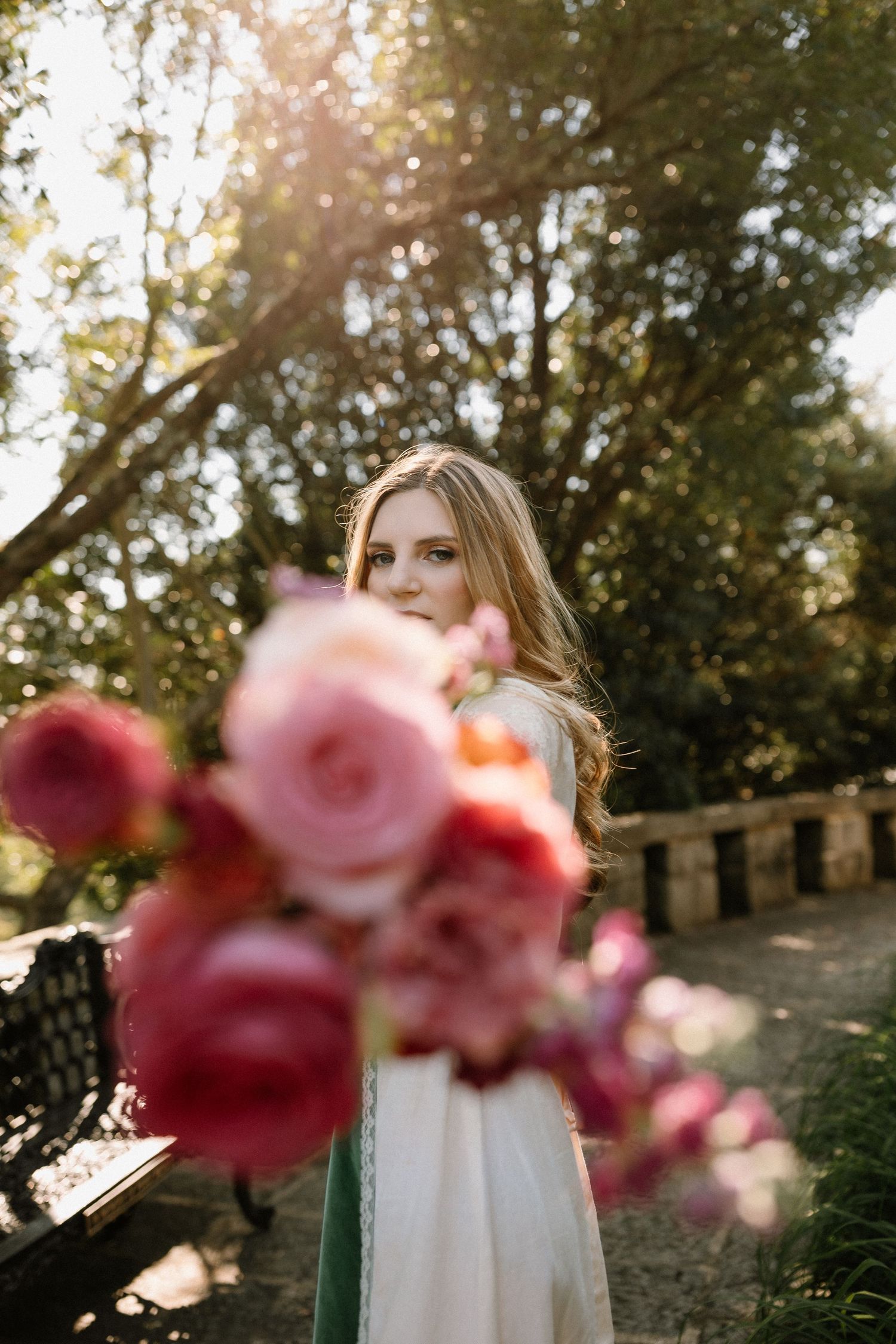 A young woman in a white dress holds a bouquet of pink and red roses outdoors in a sunlit garden.