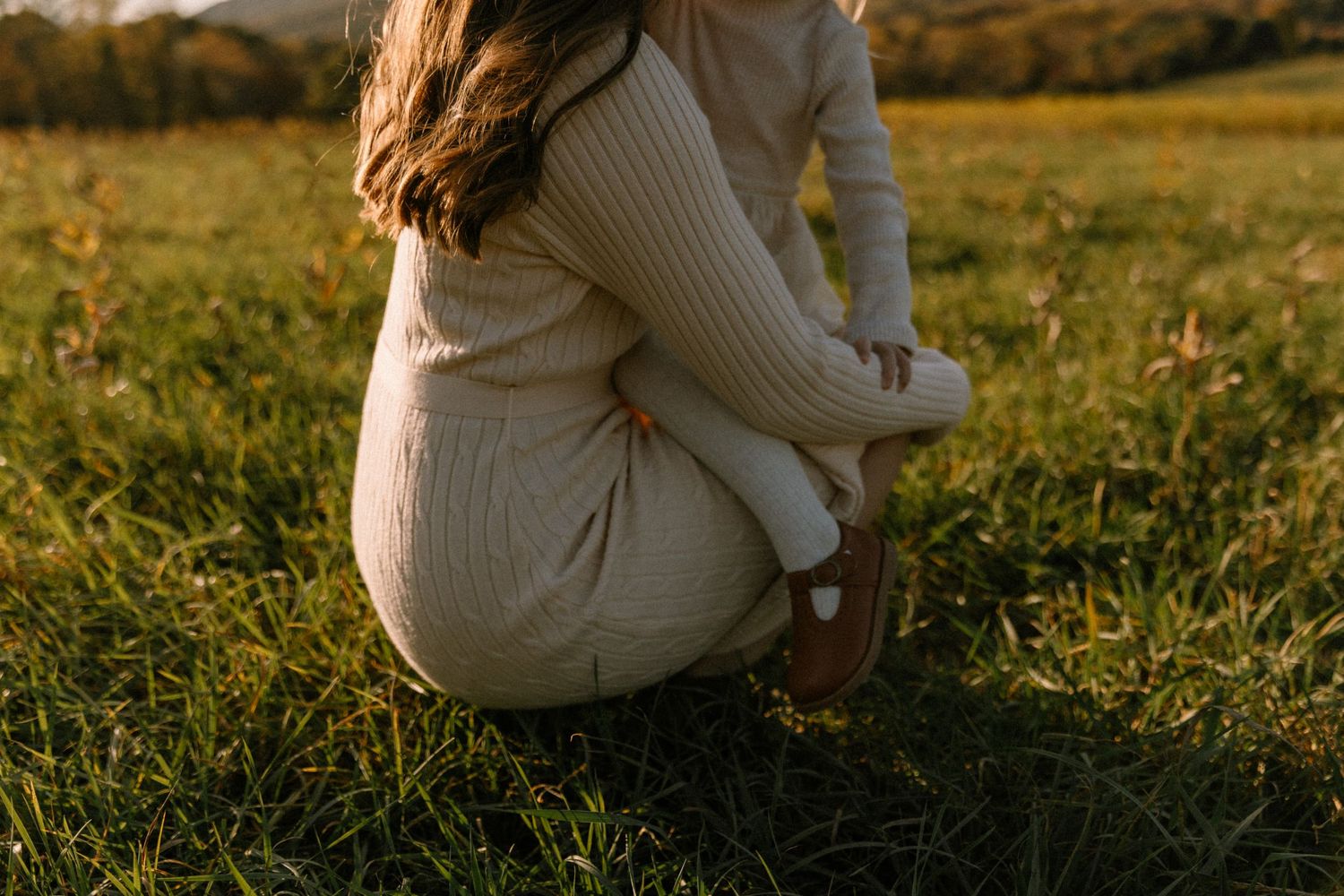 Woman in cream ribbed knit outfit crouching in a sunlit green meadow during golden hour.