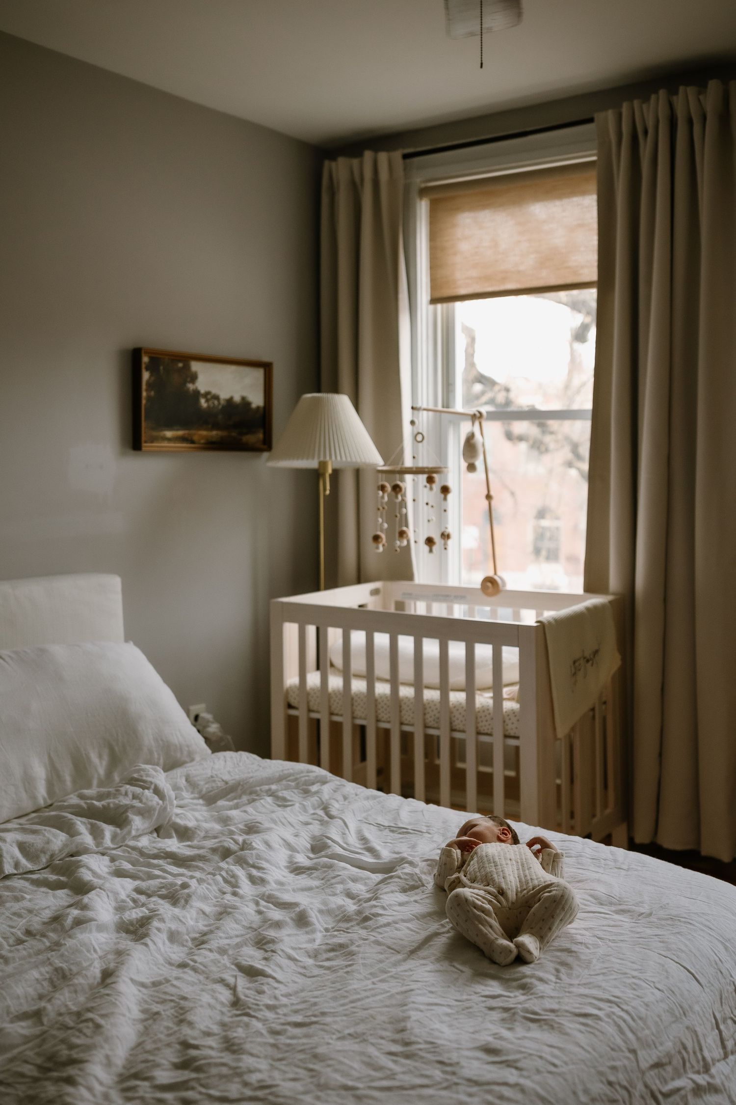 A newborn baby rests on a white bed beside a wooden crib with a mobile near a sunlit window in a cozy nursery.