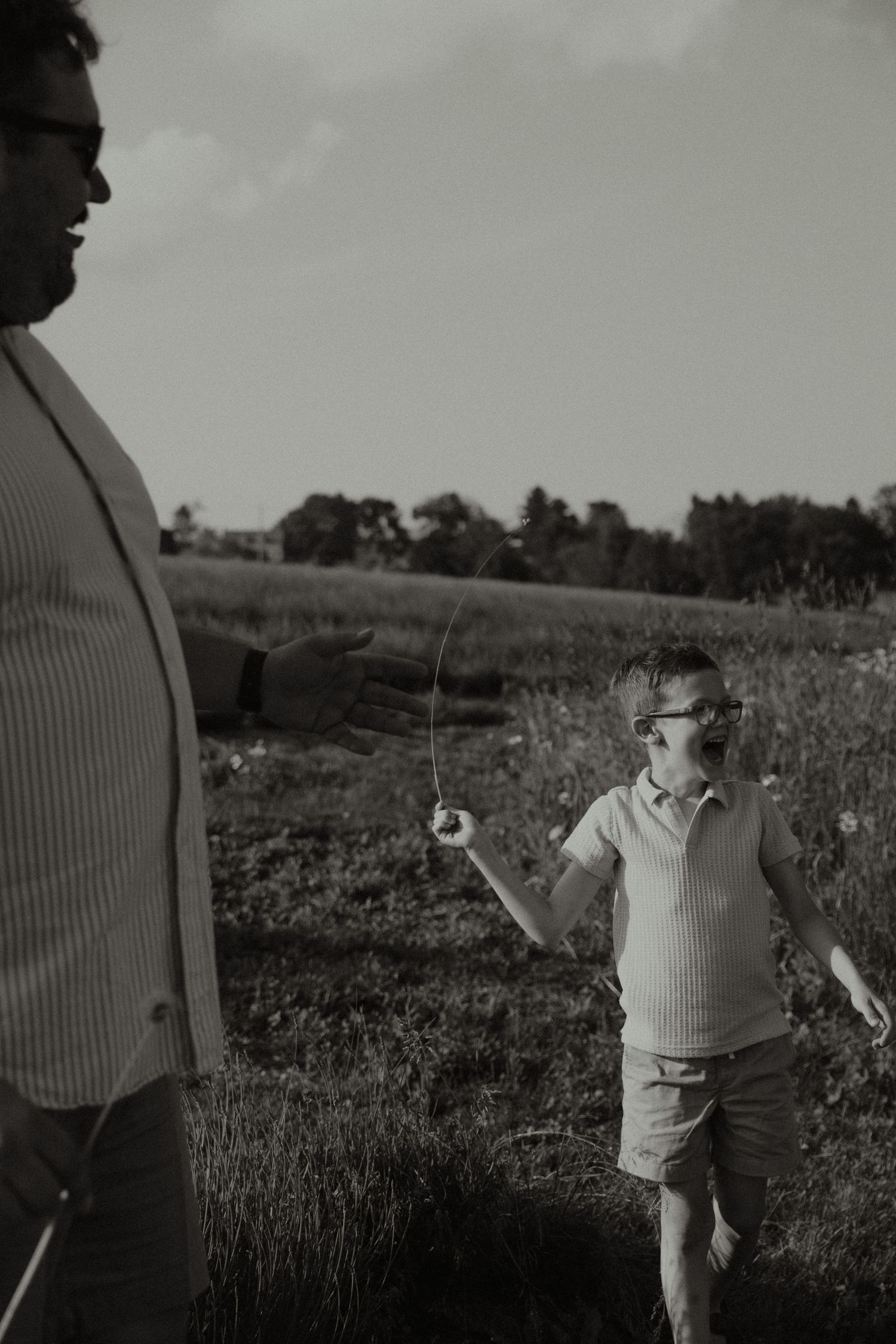 Black and white photo of a boy wearing glasses holding a fishing rod in a field with an adult nearby.