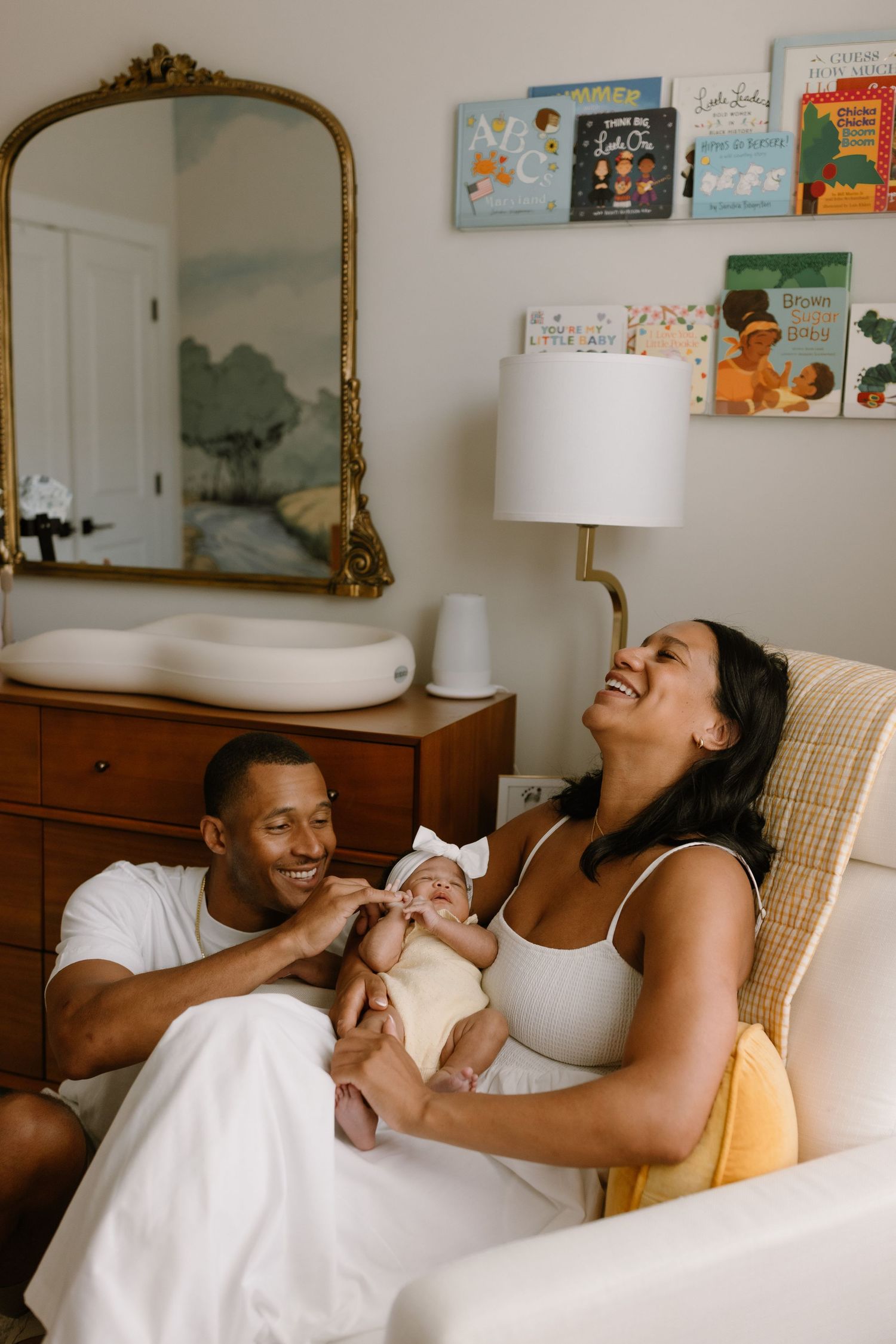 Happy couple sitting in a nursery chair, smiling and holding their newborn baby, with children's books displayed on the wall.