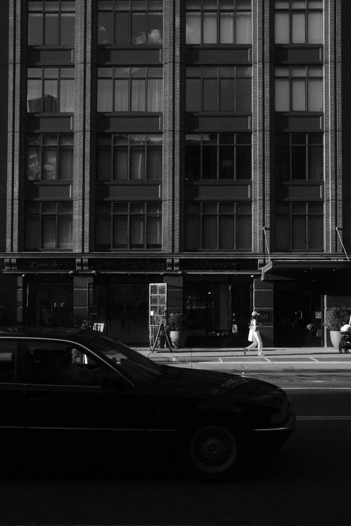 Black and white urban street scene with a car passing a large building, pedestrian walking on sidewalk in sunlight.