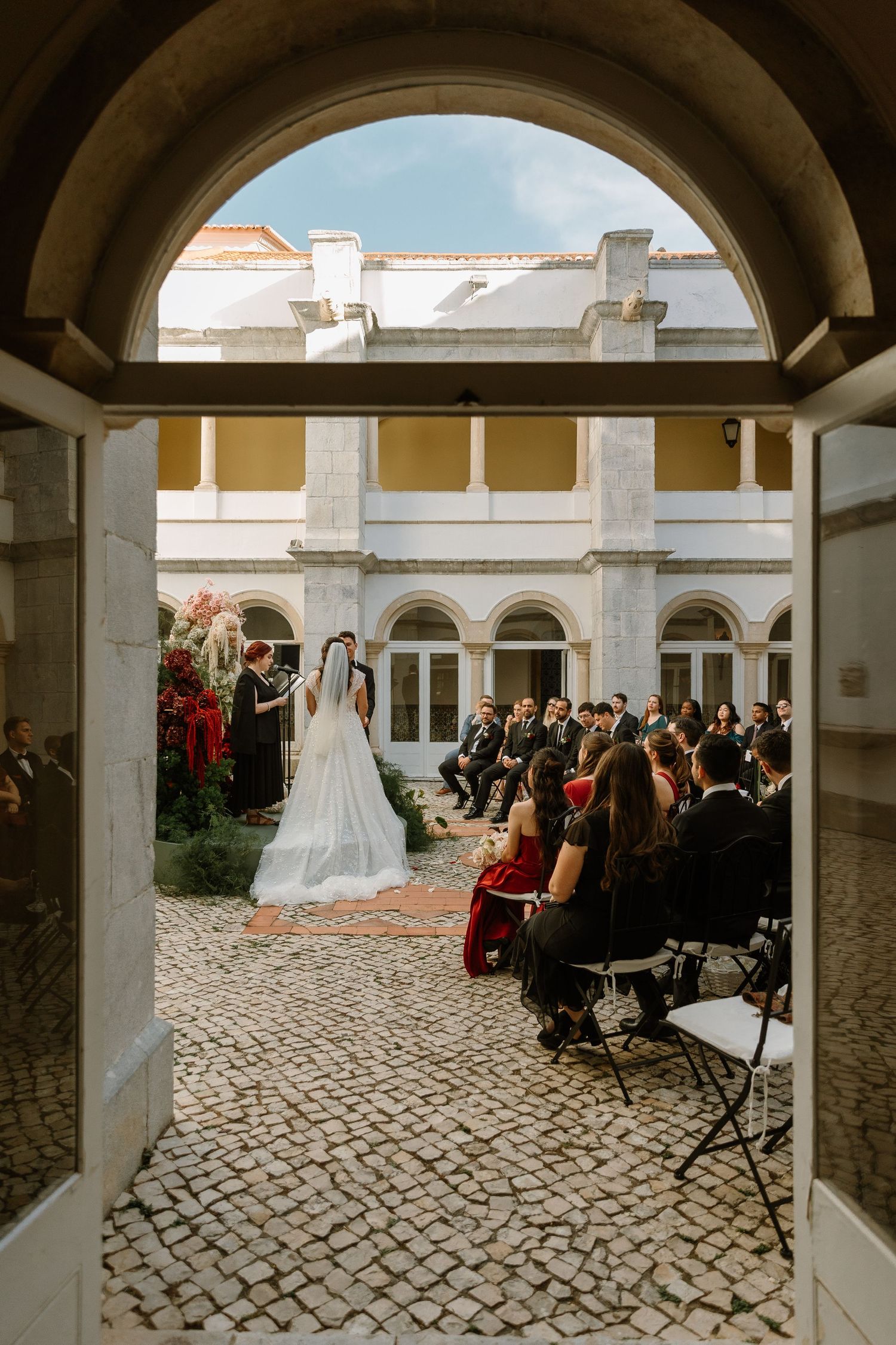 Outdoor wedding ceremony in elegant courtyard with cobblestone floor, bride in white gown, groom, and seated guests.