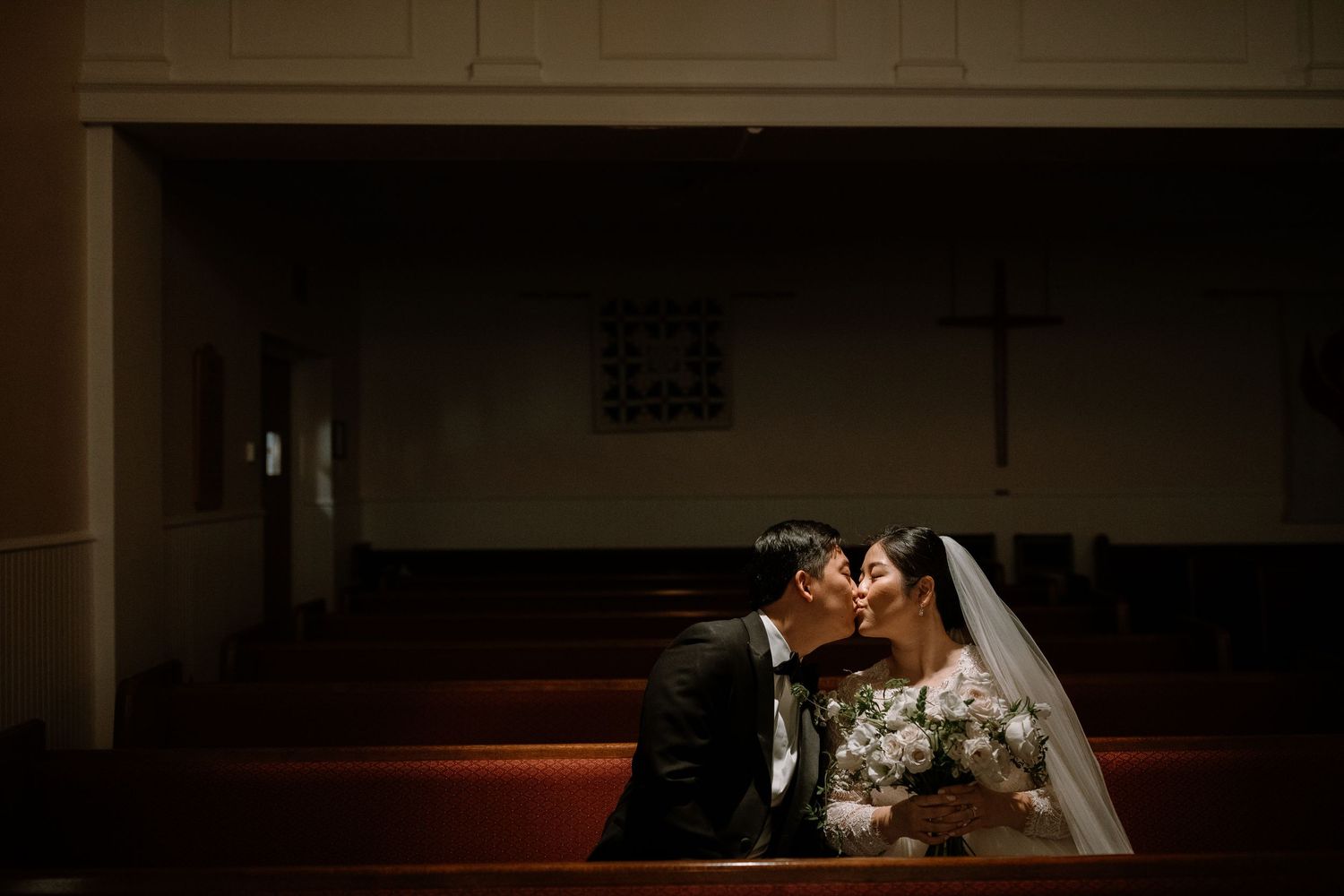 A bride and groom share a romantic kiss while seated in a church pew, with a cross visible in the background.