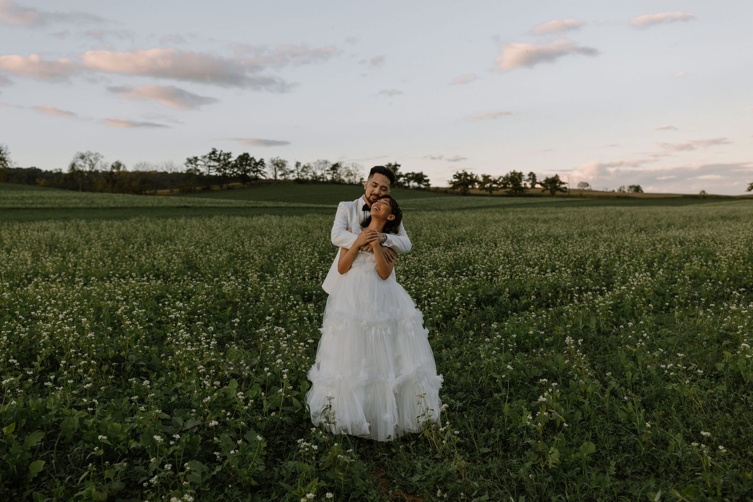 Bride and groom embrace in a lush green field at sunset, wearing elegant wedding attire in a romantic outdoor ceremony.