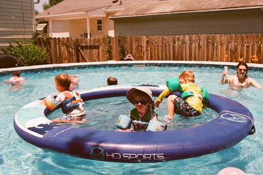 Children playing in an HD Sports inflatable ring float in a backyard swimming pool on a sunny day.